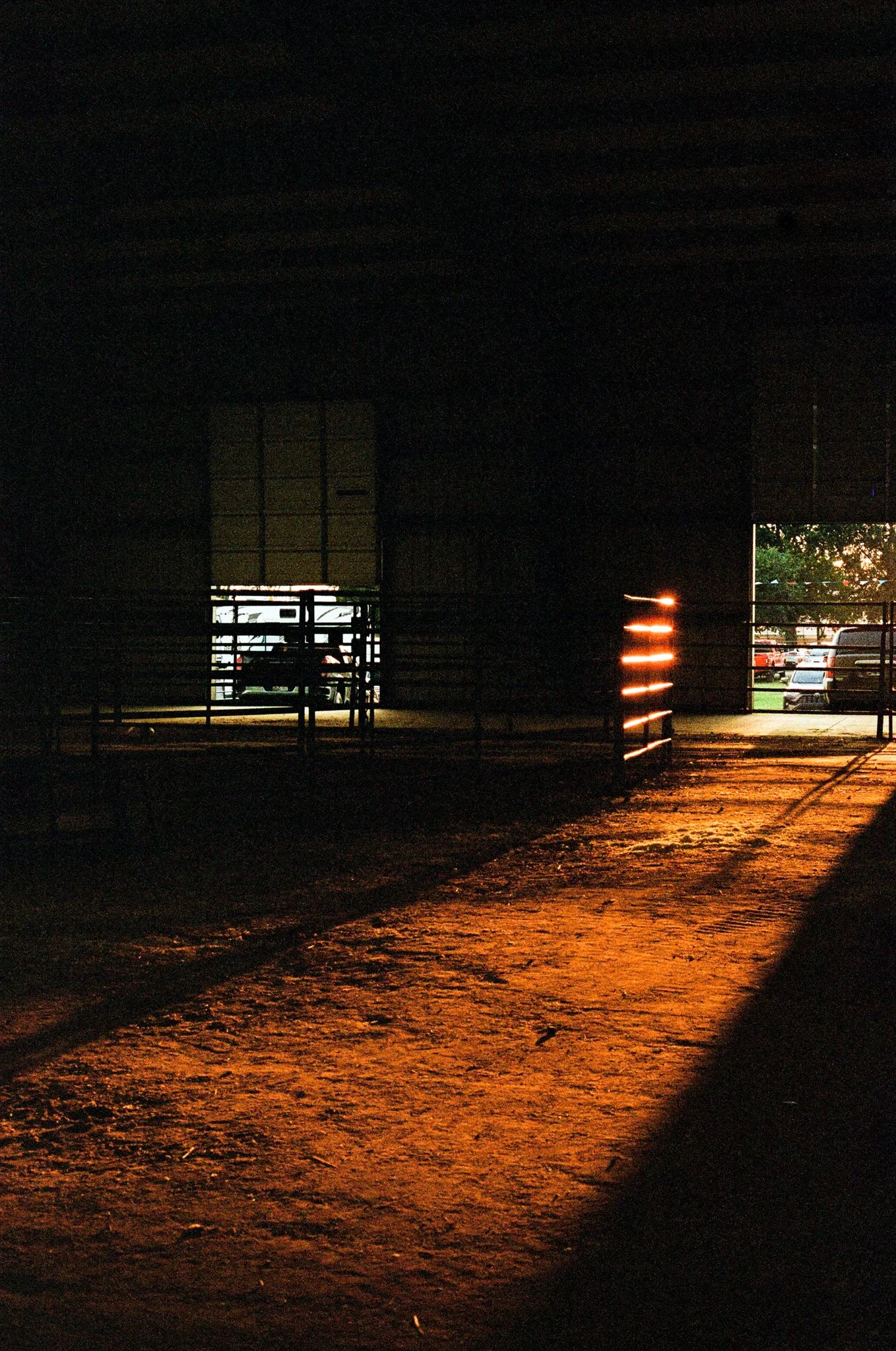 A dark interior space with a small opening revealing a parking lot with cars and trees outside. The ground is illuminated with warm, orange lighting reflecting on the surface.