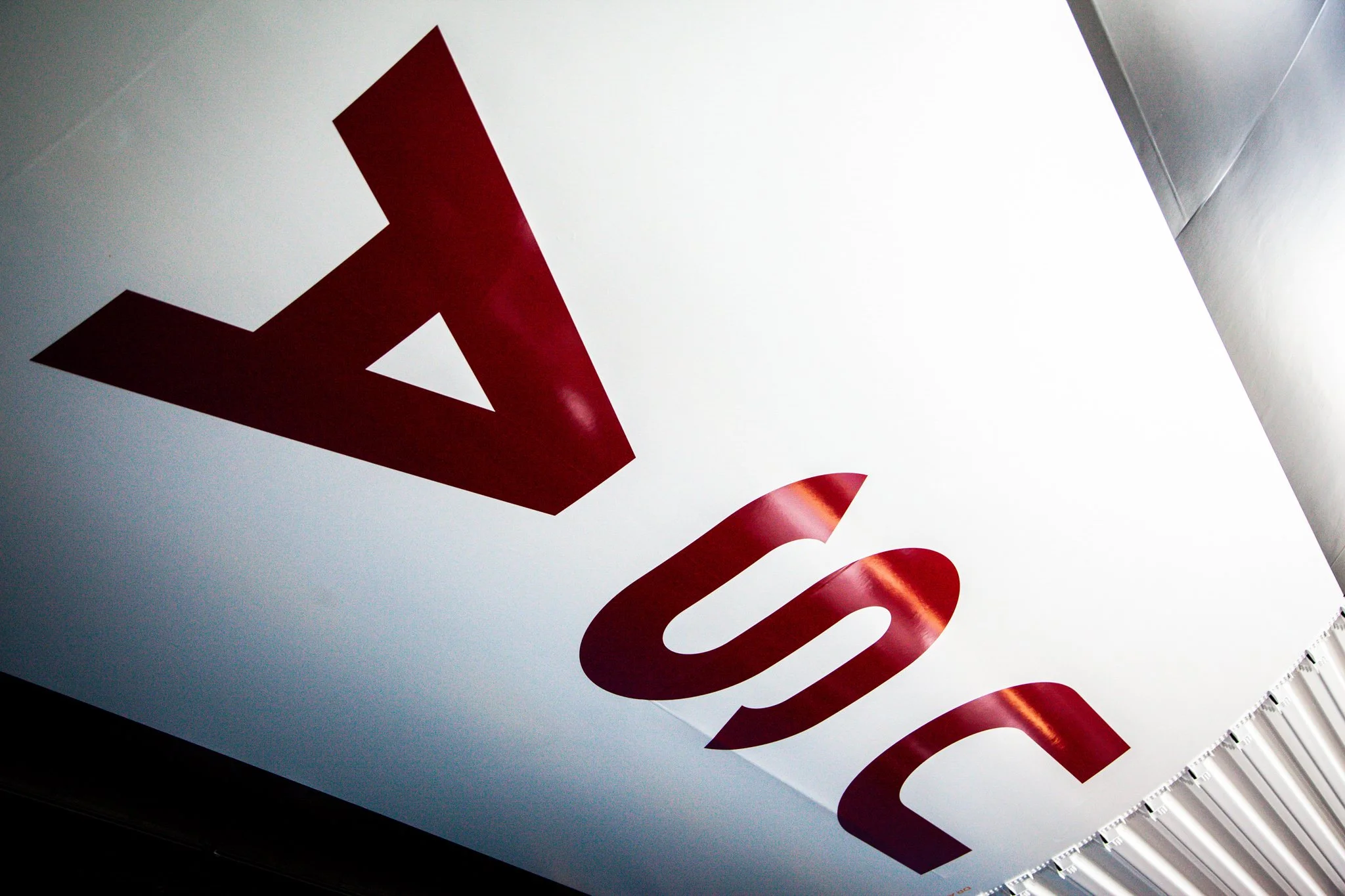 Close-up of a sign with red letters on a white background, taken at an angle, with part of a white ceiling and a row of fluorescent lights visible.