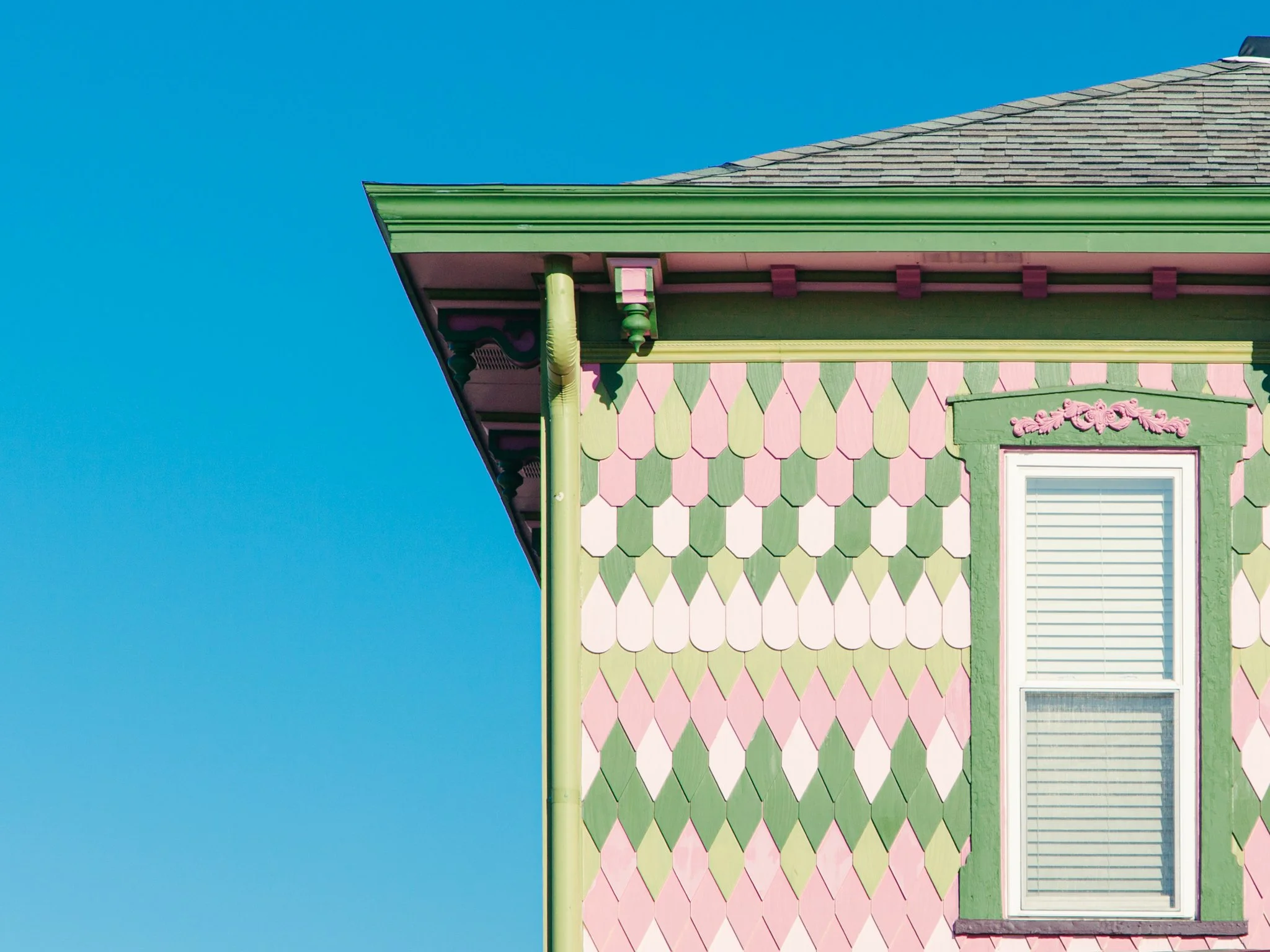 Close-up of a colorful house corner with pink, green, and purple scalloped shingles, green trim, and a white window with blinds against a blue sky.