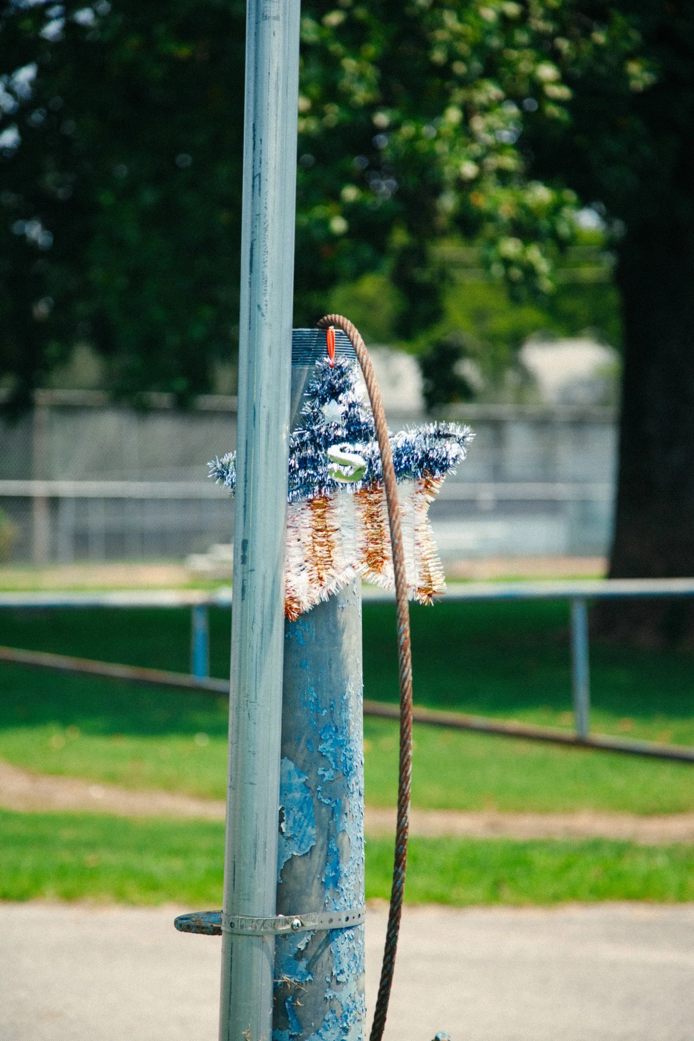 A weathered metal pole with chipped paint, holding a basket with a 4th of July wreath decoration, connected to a climbing rope, set in a park with green grass and trees in the background in Rockvale, TN