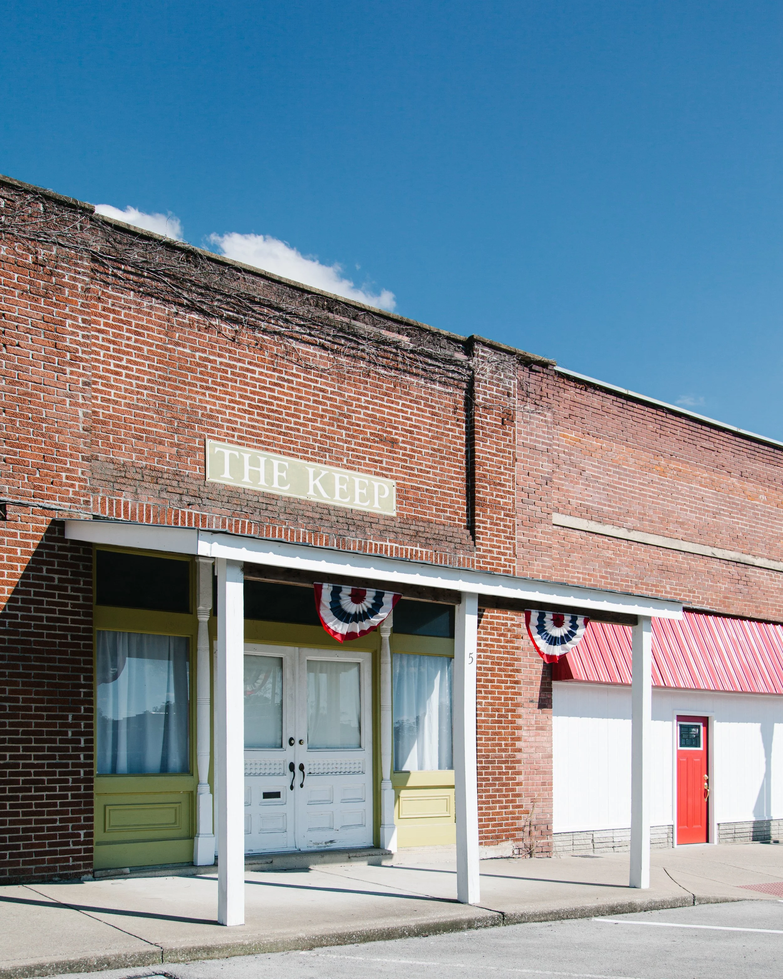 The exterior of a brick building with three storefronts, each with a different colored door and patriotic bunting. The upper part of the building has a sign that reads "THE KEEP." The sidewalk in front is empty, and the sky above is bright blue with 