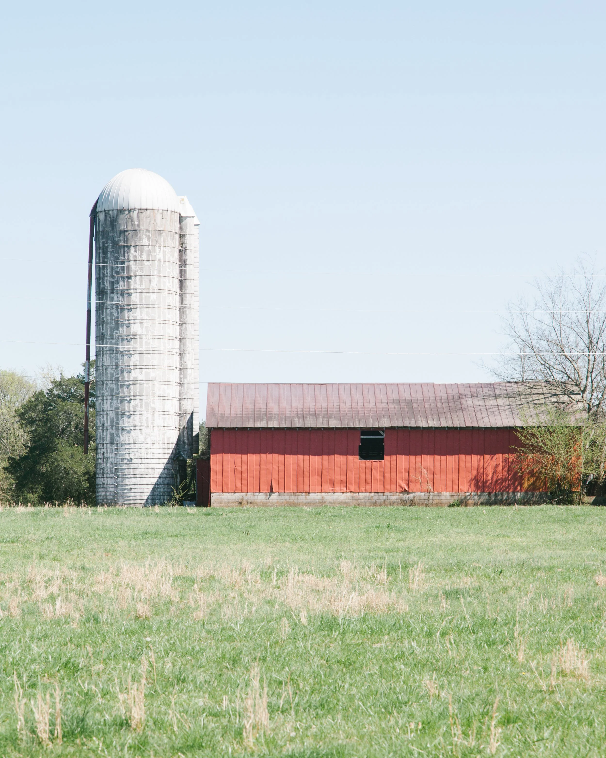 A rural farm scene with a tall white silo, a red barn with a rusted metal roof, and a grassy field in the foreground. Trees are visible in the background under a clear blue sky.