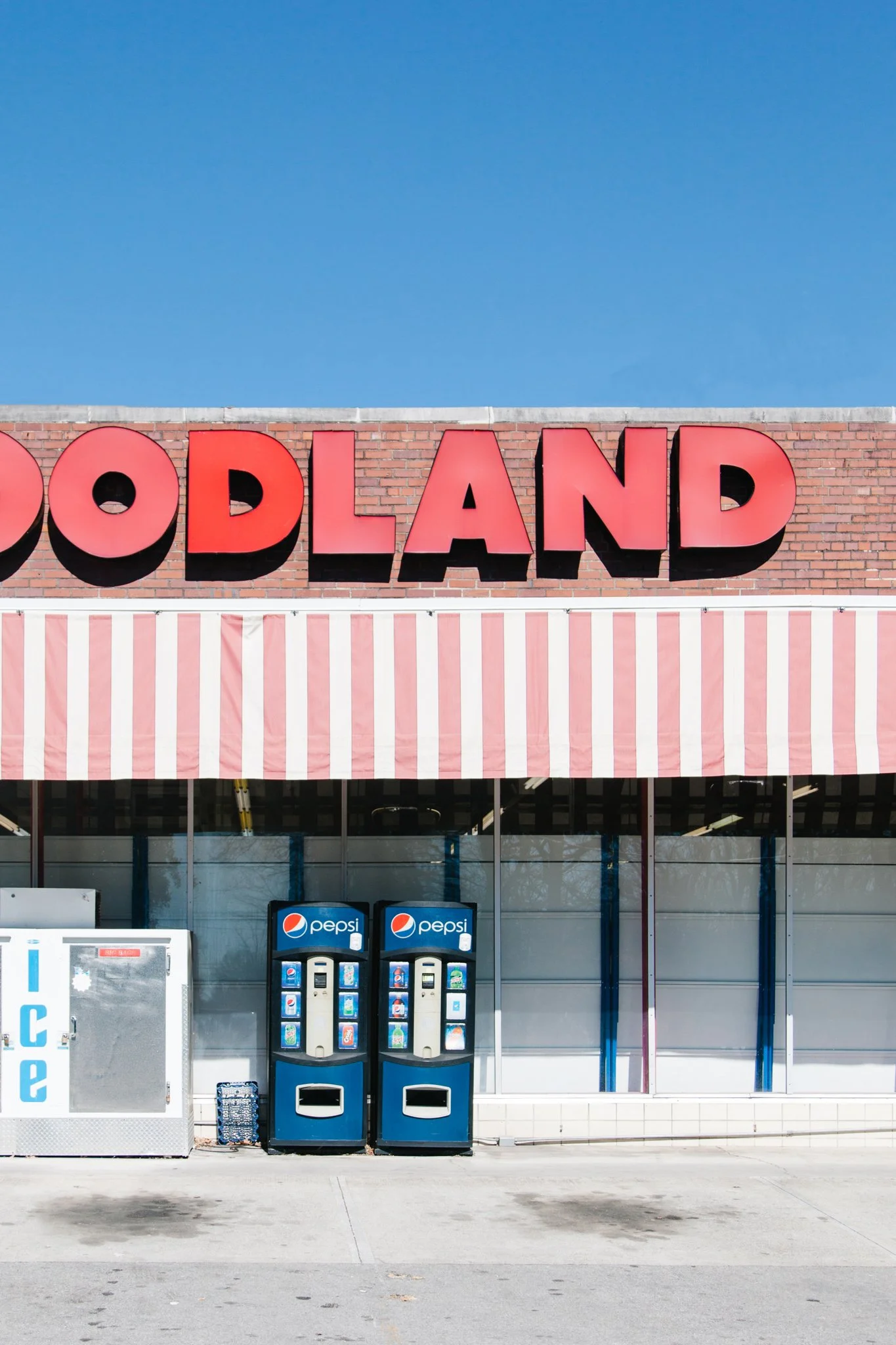 Storefront with large red letters spelling 'ODLAND' on a brick wall, pink and white striped awning, and vending machines outside, under a clear blue sky.