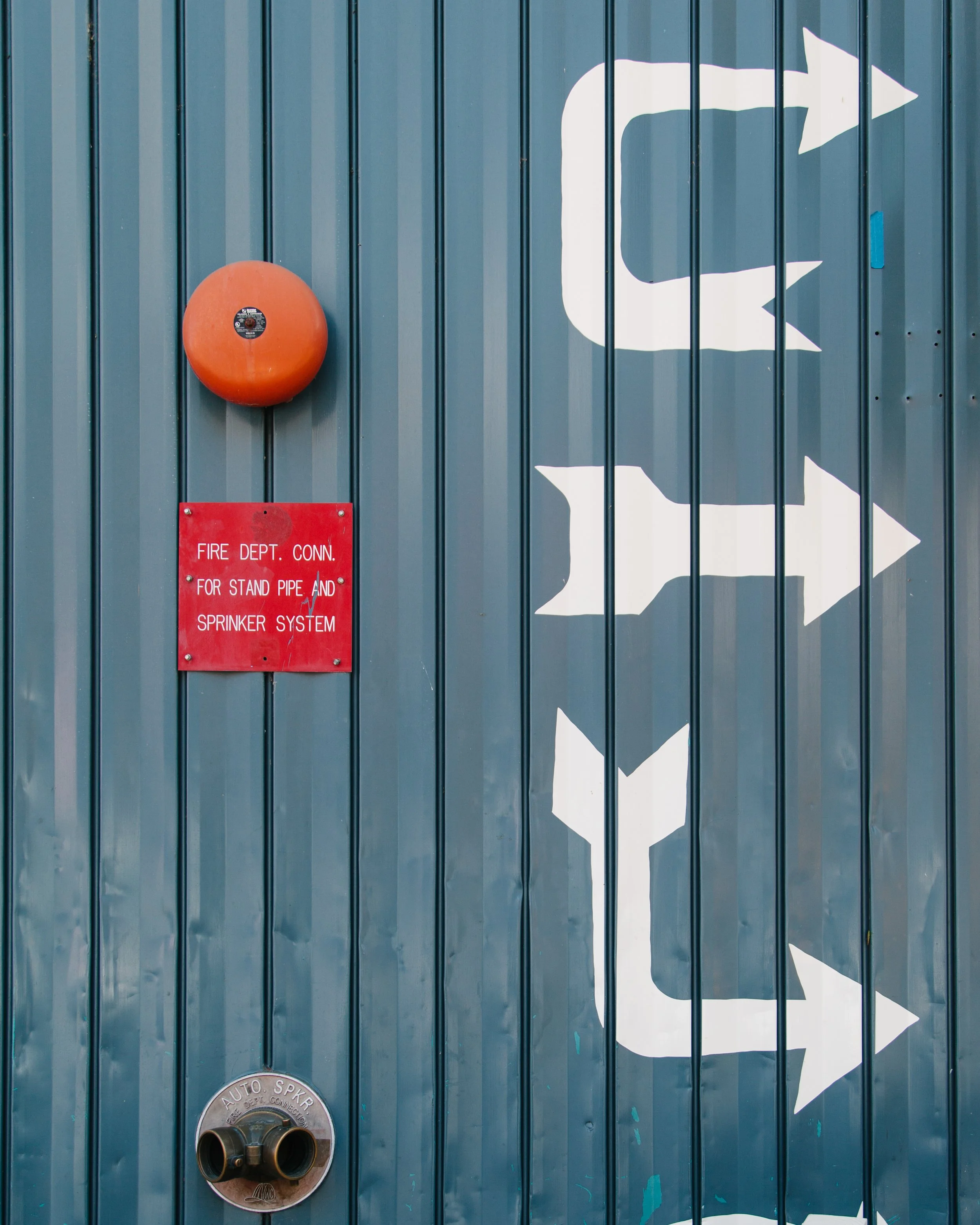 Blue metal wall with a large white arrow pointing to the right, a red sign indicating fire department connection for standpipe and sprinkler system, an orange fire alarm bell, and a metal fire department connection with two large hoses.