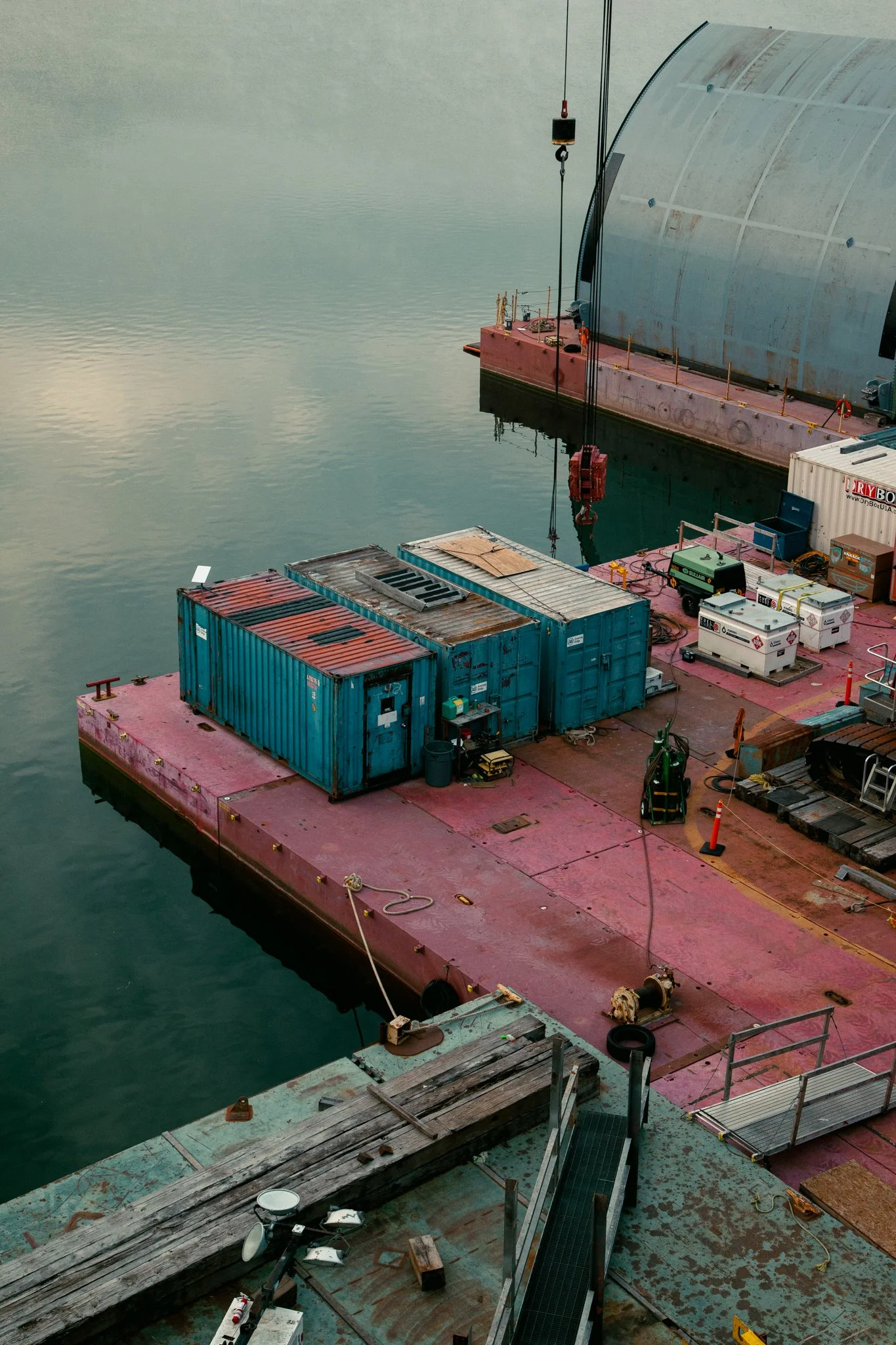 A floating dock with containers, equipment, and a large curved metal structure in a harbor.