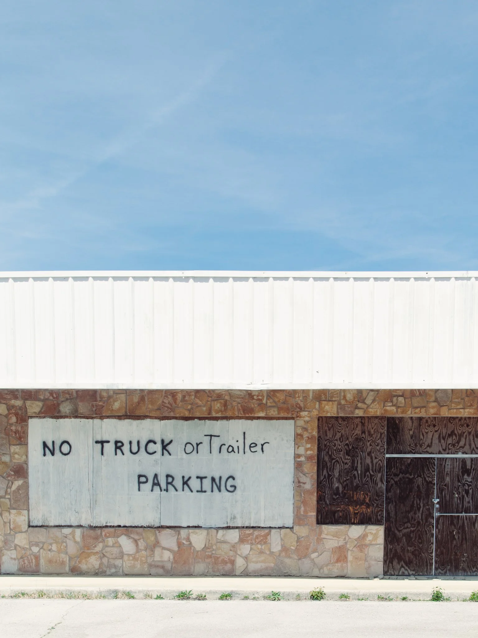 A building with a stone facade and a white, corrugated metal upper wall. A handwritten sign on a white board reads 'NO TRUCK or Trailer PARKING' in black paint.