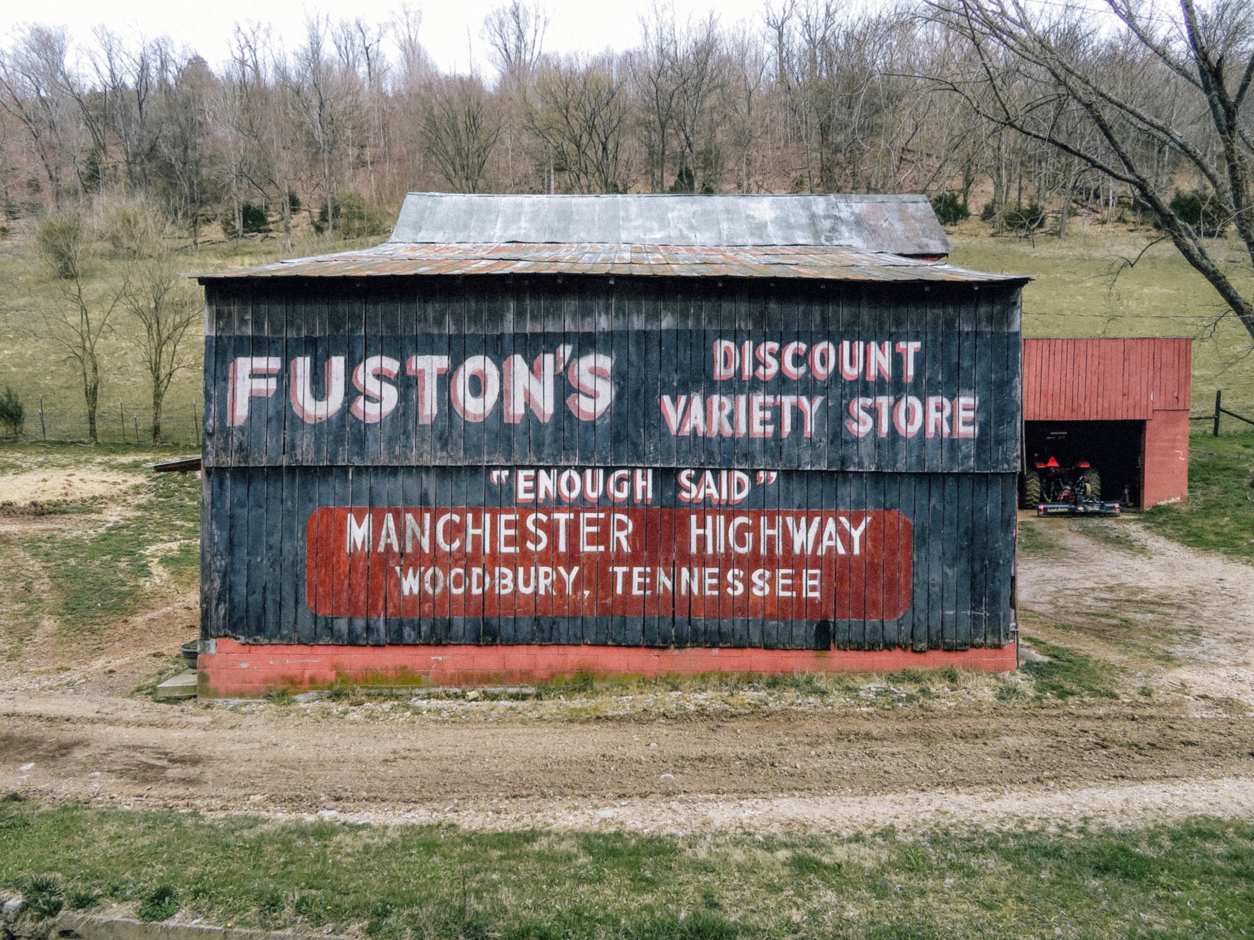 An old weathered barn with faded lettering on its side reading 'FUSTON'S DISCOUNT VARIETY STORE.' The barn is situated in a rural area with leafless trees in the background, a grassy field, and a dirt path in the foreground, under an overcast sky.