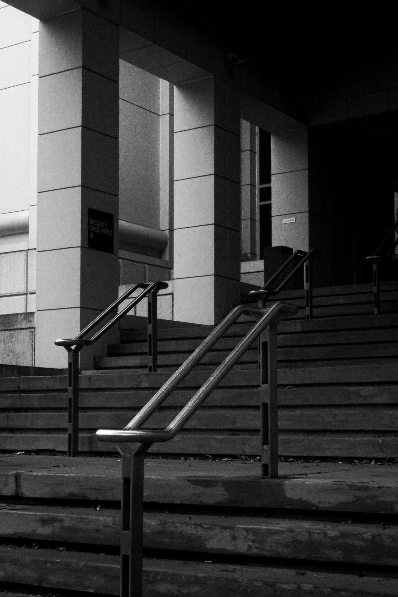 Black and white photo of a building entrance with stairs, metal handrails, and a sign that reads 'Security Checkpoint'.