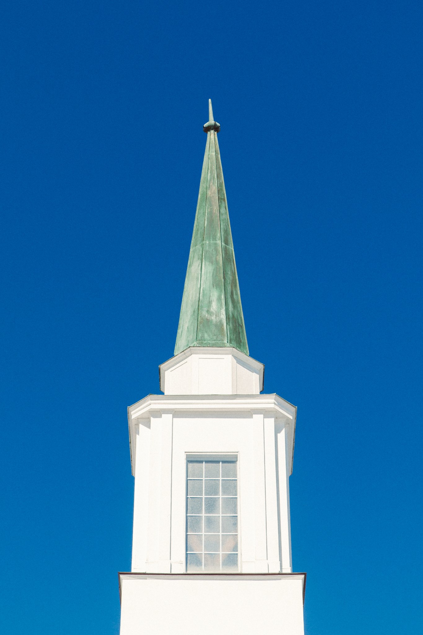 A white church steeple with a clock in the middle, topped with a greenish copper spire, against a bright blue sky.