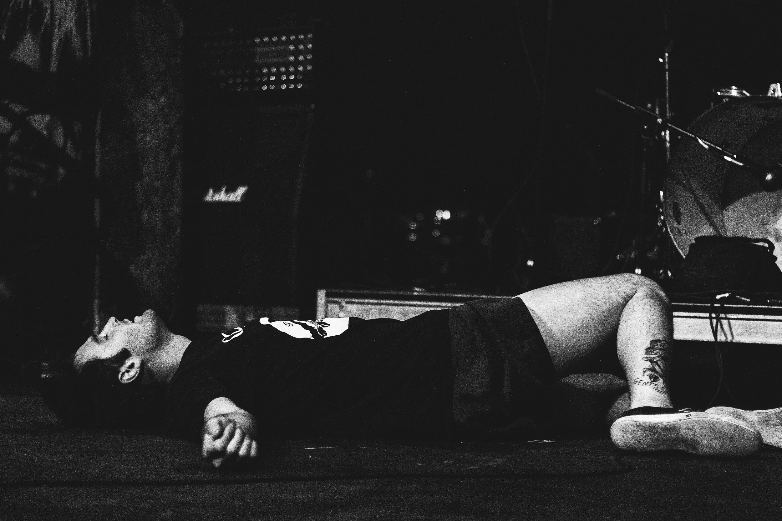 A person lies on the floor with their eyes closed, wearing a dark shirt with a graphic design, and sneakers. The room contains musical equipment including a Marshall amplifier, a drum set, and a microphone stand, suggesting a music studio or rehearsa