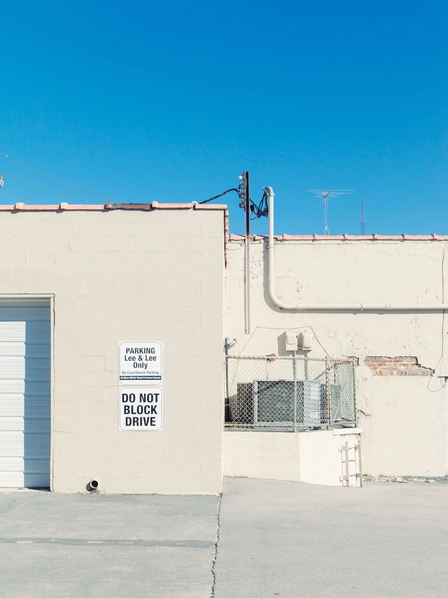 A beige building with a pink tiled roof, a garage door, and a sign that says 'Parking Lee & Lee Only' and 'Do Not Block Drive.' There are some wires, antennas, and a fenced area on the roof, and a drainpipe running down the wall.