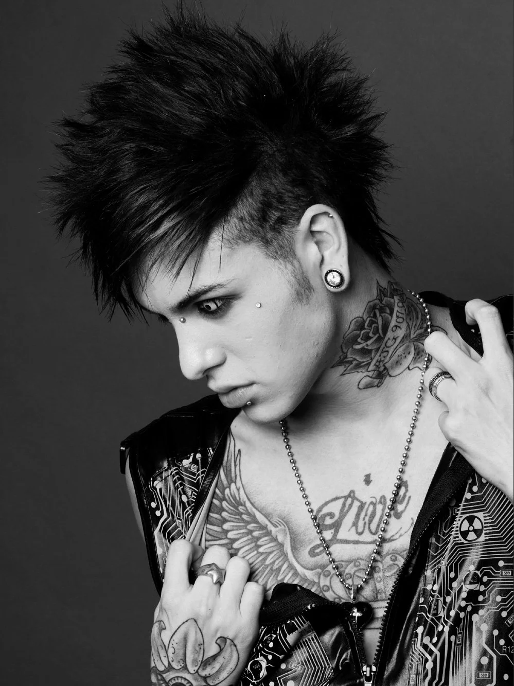 A person with spiky dark hair, tattoos, piercings, and wearing a circuit board patterned jacket, looking downward in a black-and-white portrait.