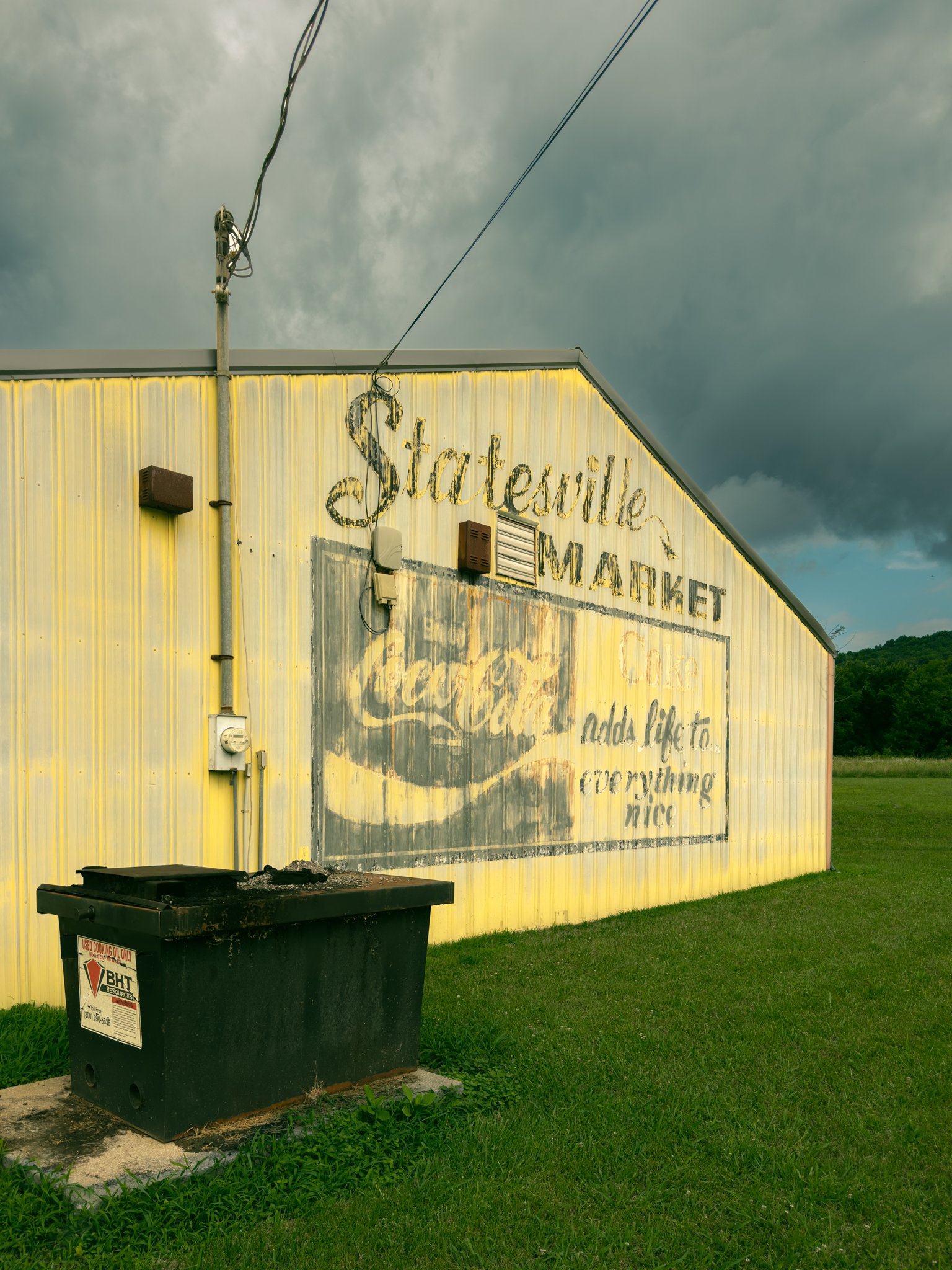 Photo of the side of a yellow building with a distressed sign that reads 'Statesville Market'. A green grass lawn and a black trash container in the foreground. Overcast sky with dark clouds in the background.