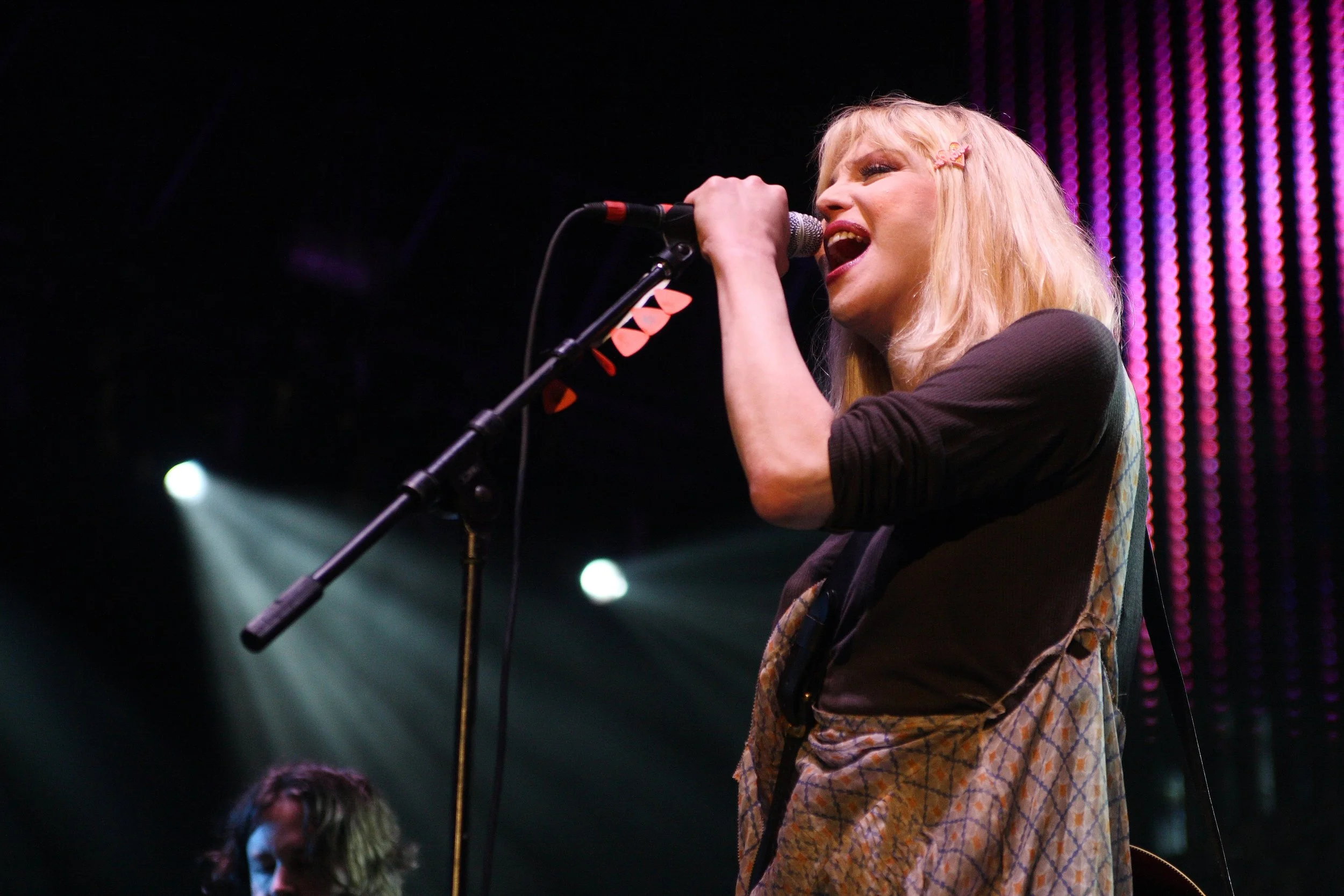A woman with blonde hair, pink hair clip, and colorful patchwork dress singing into a microphone on stage with purple and pink lighting in the background.