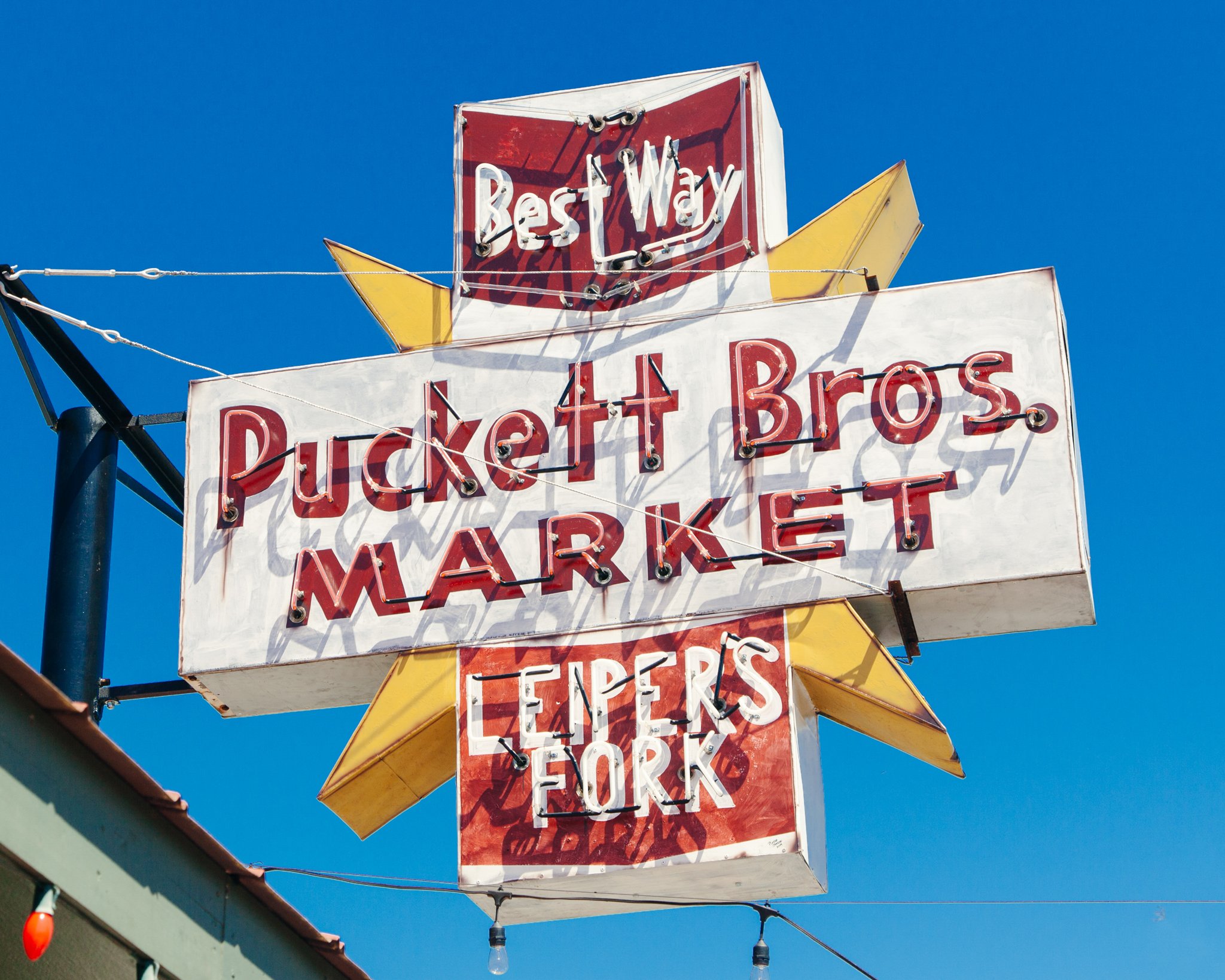 Vintage neon signs for a restaurant or bar, including one that reads 'Best Way' and another partially visible sign with 'FURE'. The signs are mounted on a pole against a clear blue sky, and are illuminated with neon tubing.