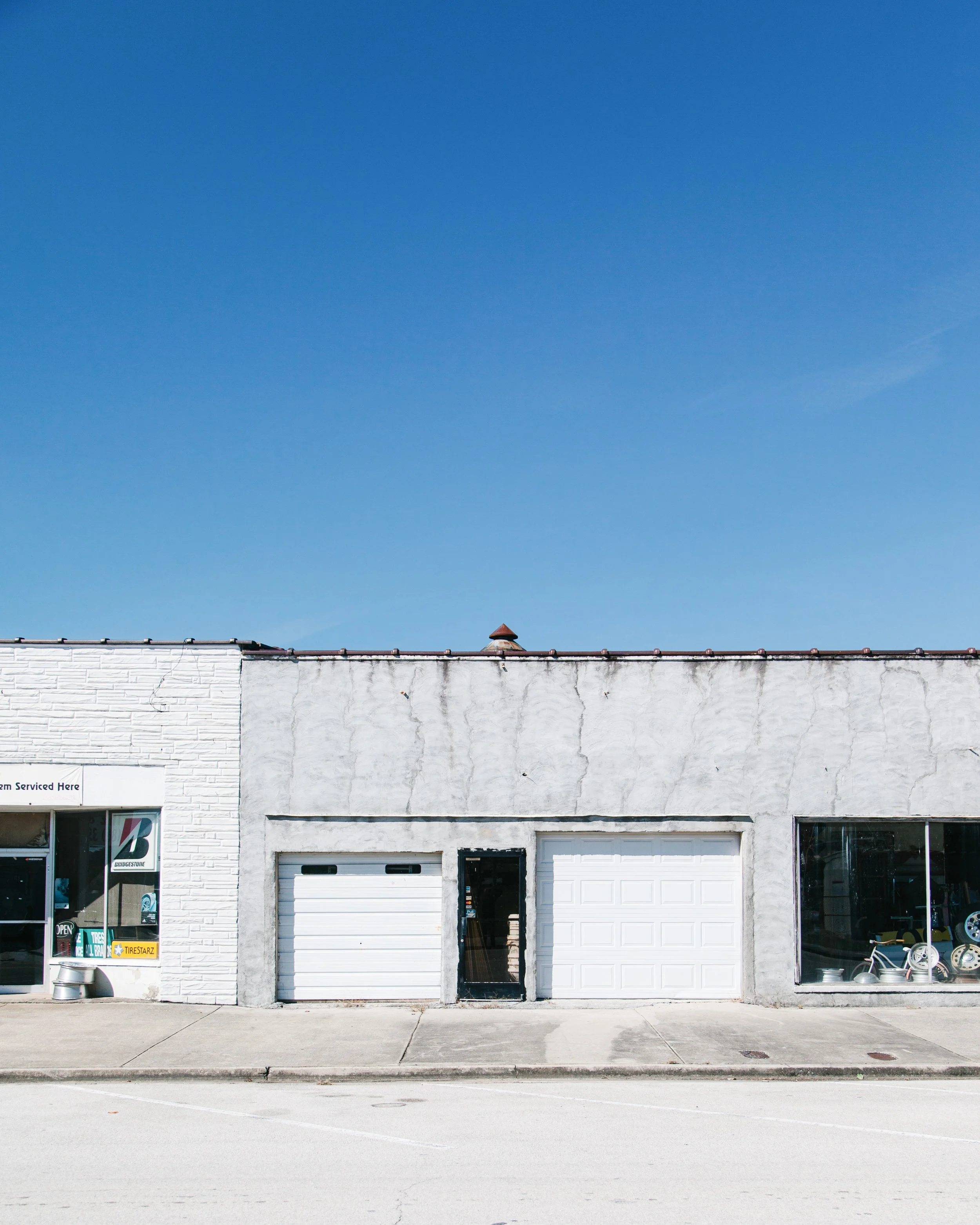 A storefront with three large garage doors, a window with car tires on display, and a sidewalk in front. The building has a white brick and stucco exterior, and a blue sky overhead.