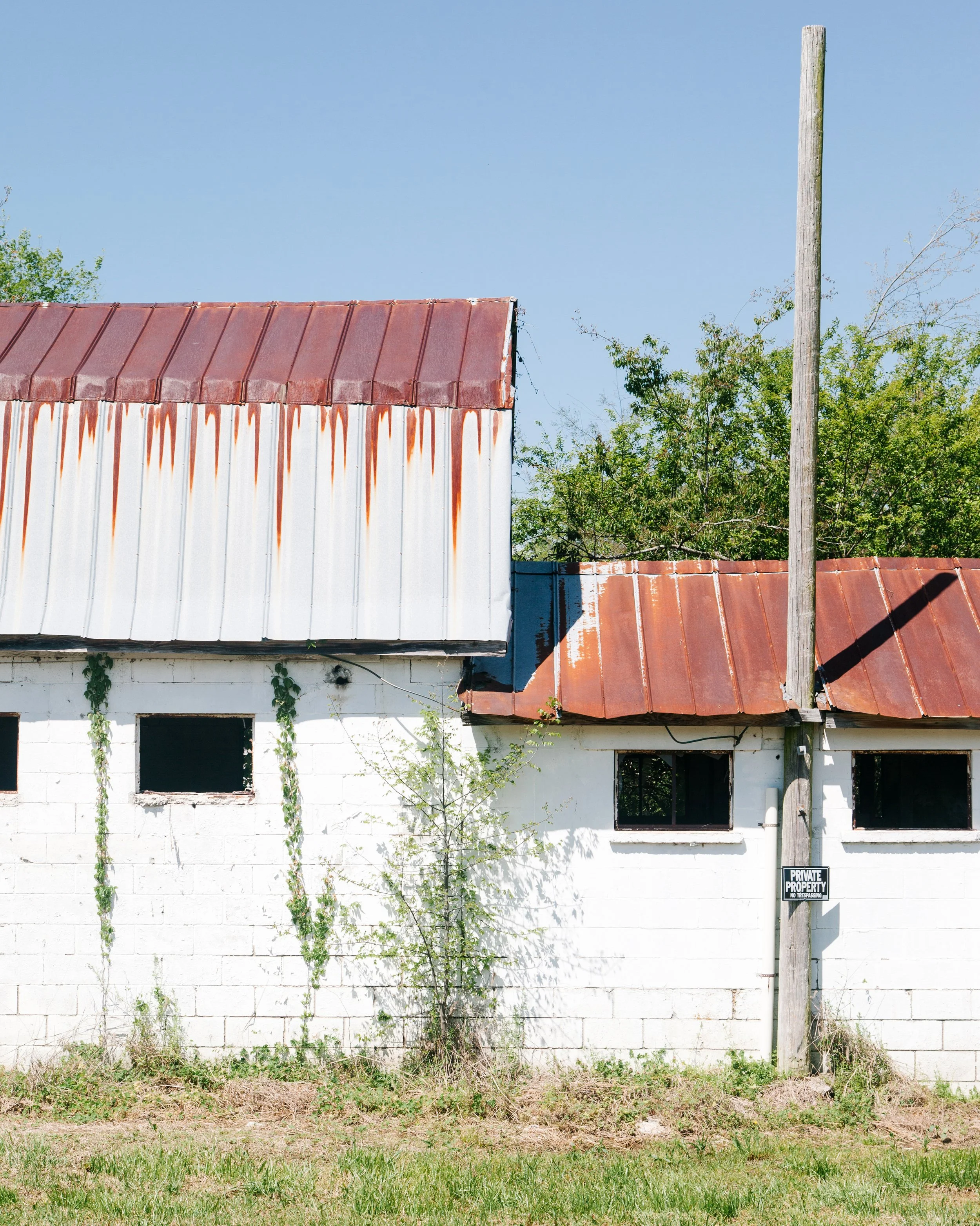 An old, weathered building with a rusted metal roof and a white brick wall, overgrown with plants and weeds, with a wooden utility pole blocking part of the view, and a sign reading "Private Property."