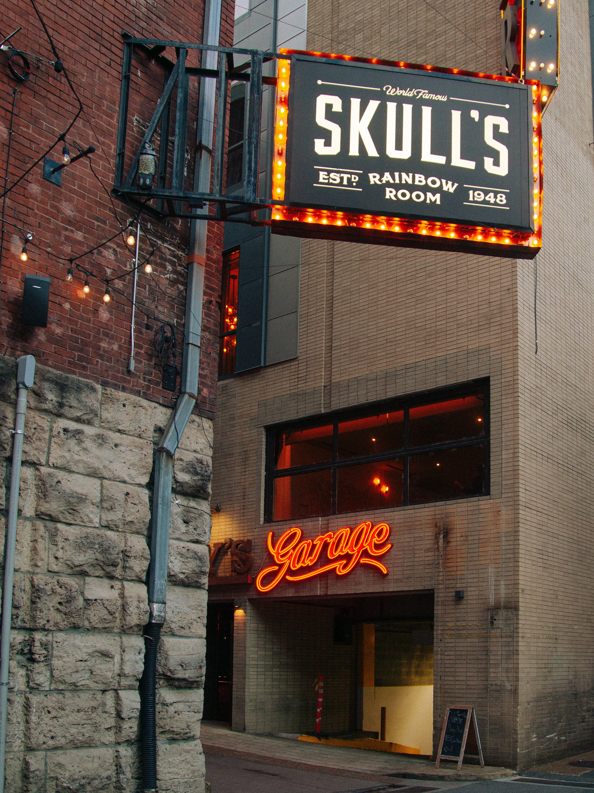 Neon sign reading 'Skull's World Famous Rainbow Room Est. 1948' on a building facade, with brick and modern exterior walls, and string lights visible on the left.