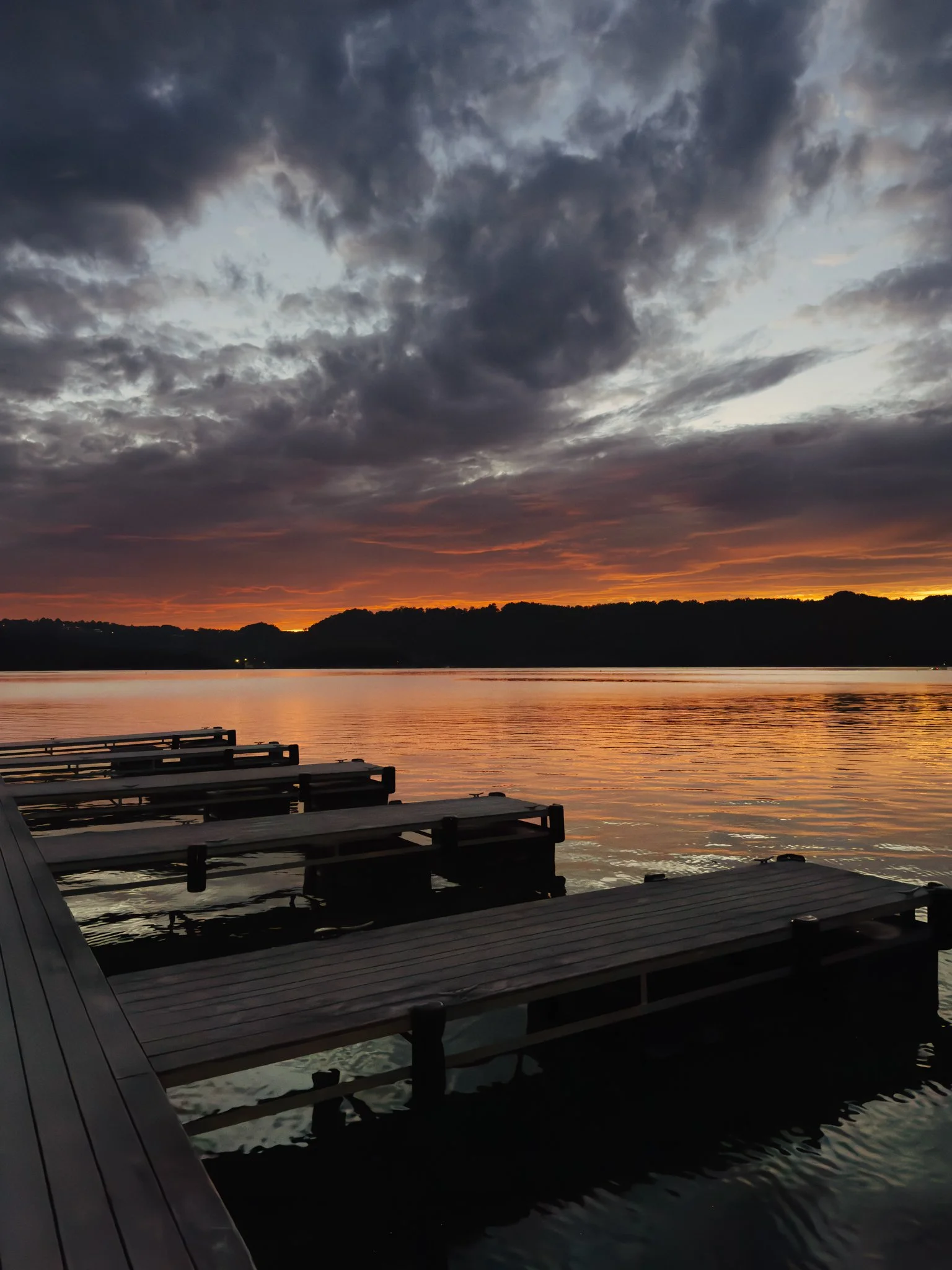 A lakeside scene during sunset with a sky filled with dark clouds and orange hues. Wooden docks extend into the water, reflecting the colors of the sky.