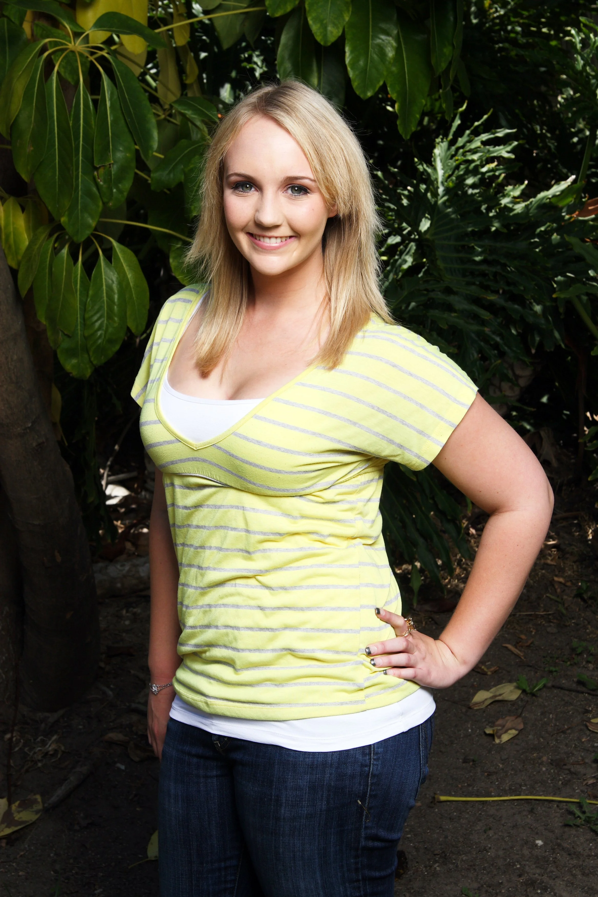 A blonde woman smiling outdoors, wearing a yellow and gray striped shirt and blue jeans, standing among large green leaves and plants.