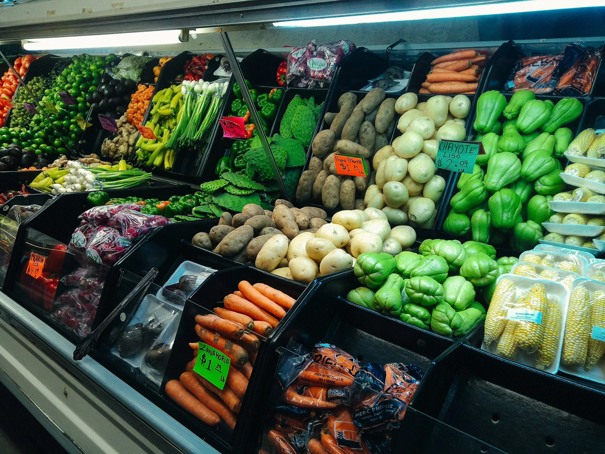 Display of fresh vegetables at a grocery store, including carrots, green bell peppers, potatoes, chayote, and sweet corn, with handwritten price tags.