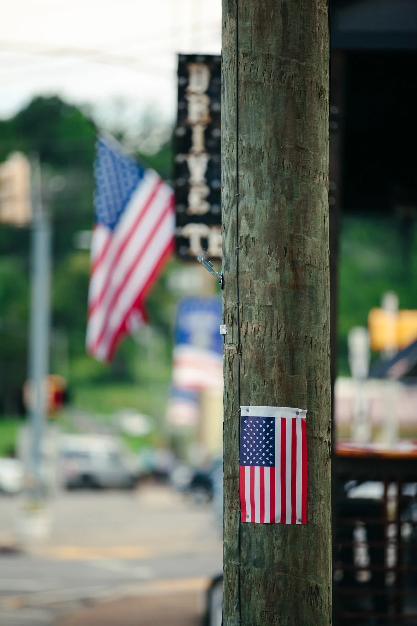 A wooden utility pole decorated with a small American flag attached vertically. Additional American flags are visible blurred in the background, possibly hanging or waving, indicating a patriotic display or celebration.