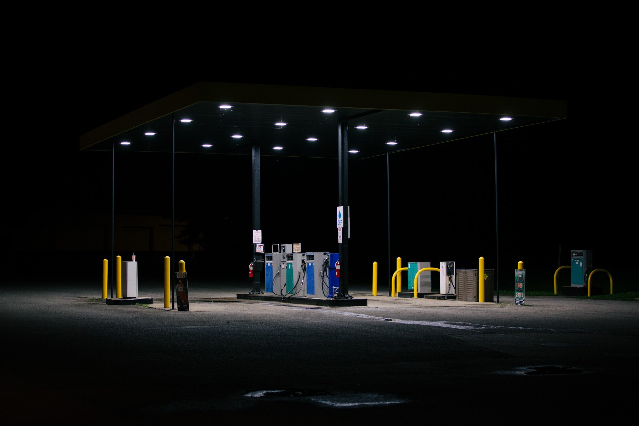 Empty gas station at night with illuminated canopy lights and fuel pumps, surrounded by yellow safety barriers.