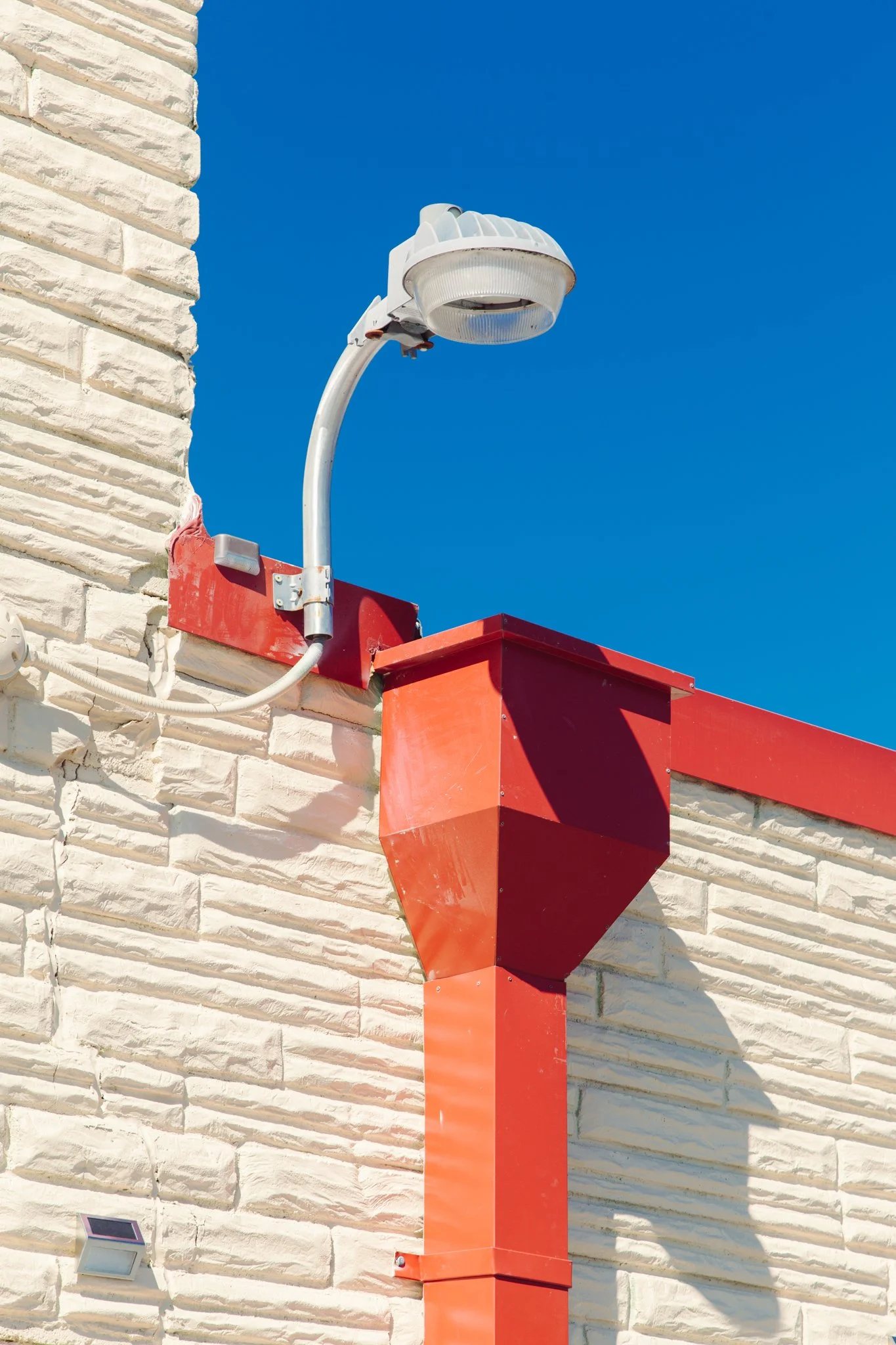 A modern streetlight mounted on a decorative brick wall against a clear blue sky, with a red vent or exhaust pipe attached to the wall below.