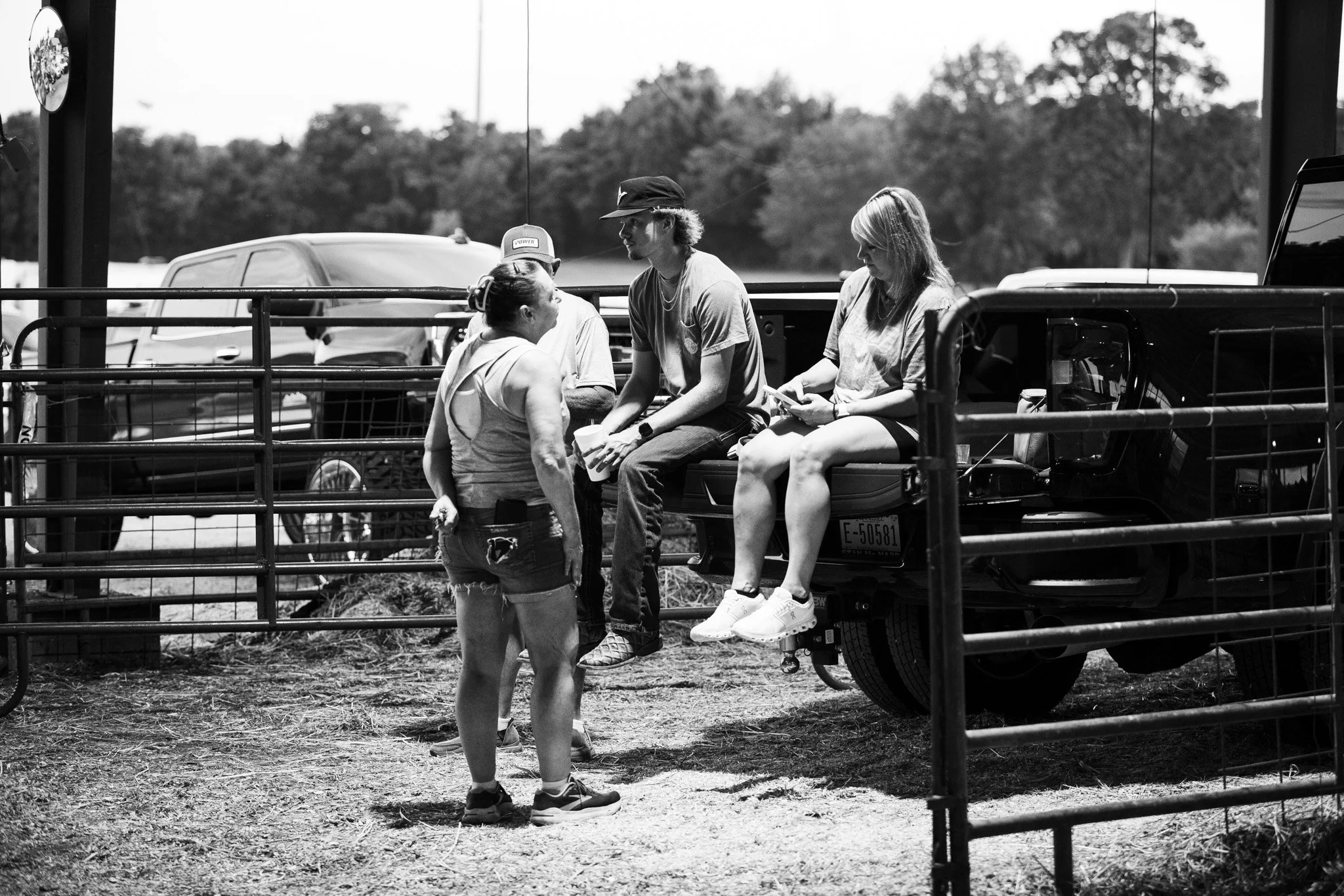 A woman with a bun talking to a young man sitting on a vehicle, with two women sitting nearby; one using a phone. There is a fence in the foreground and trees in the background.