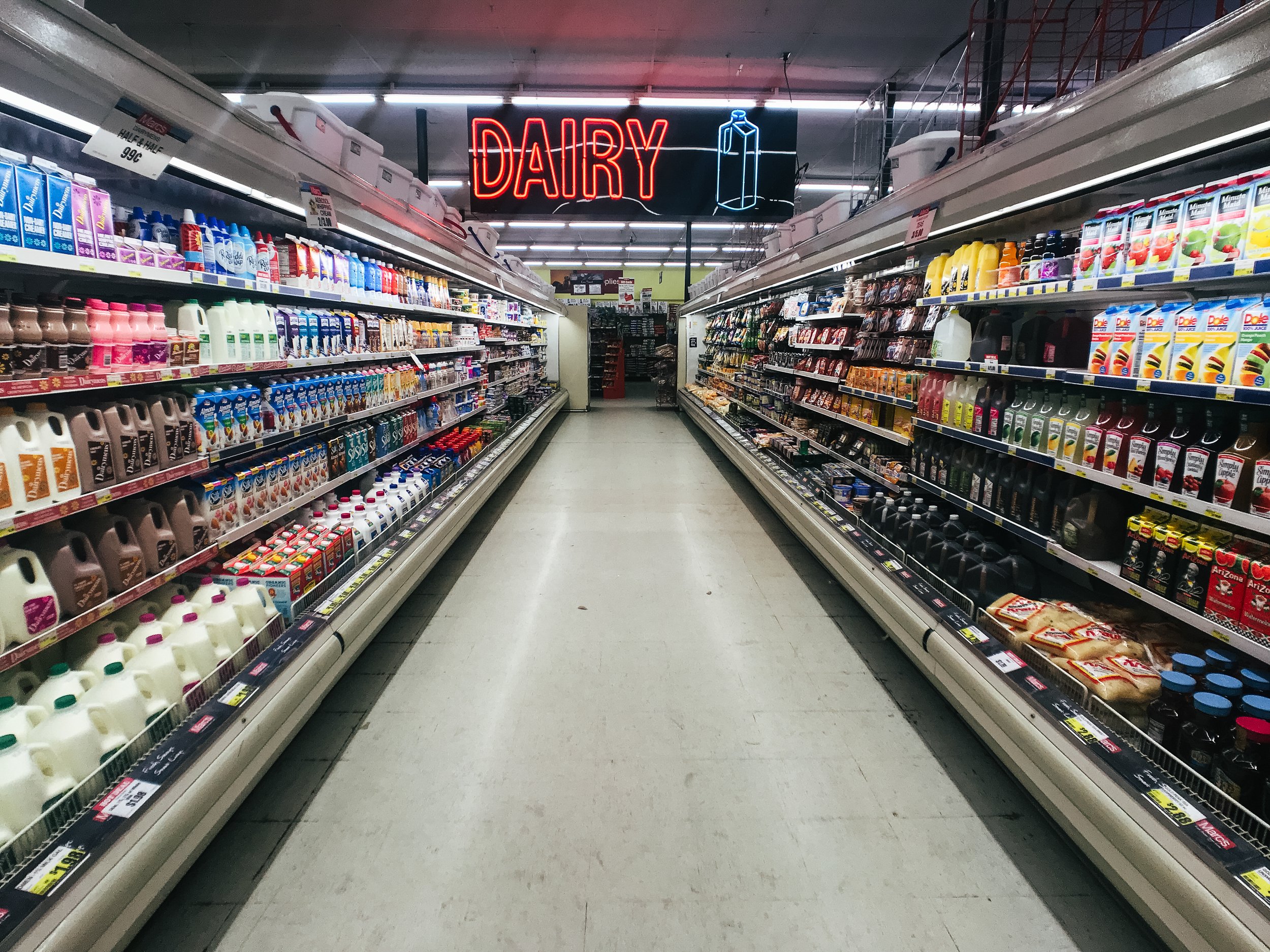 Supermarket aisle with dairy products, neon sign reading 'DAIRY' with a milk bottle icon above.