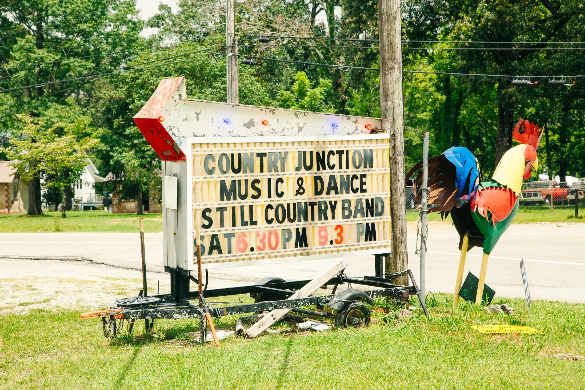 An outdoor sign advertising a country music and dance event, featuring colorful lights and a decorative rooster figure, with trees and power lines in the background.