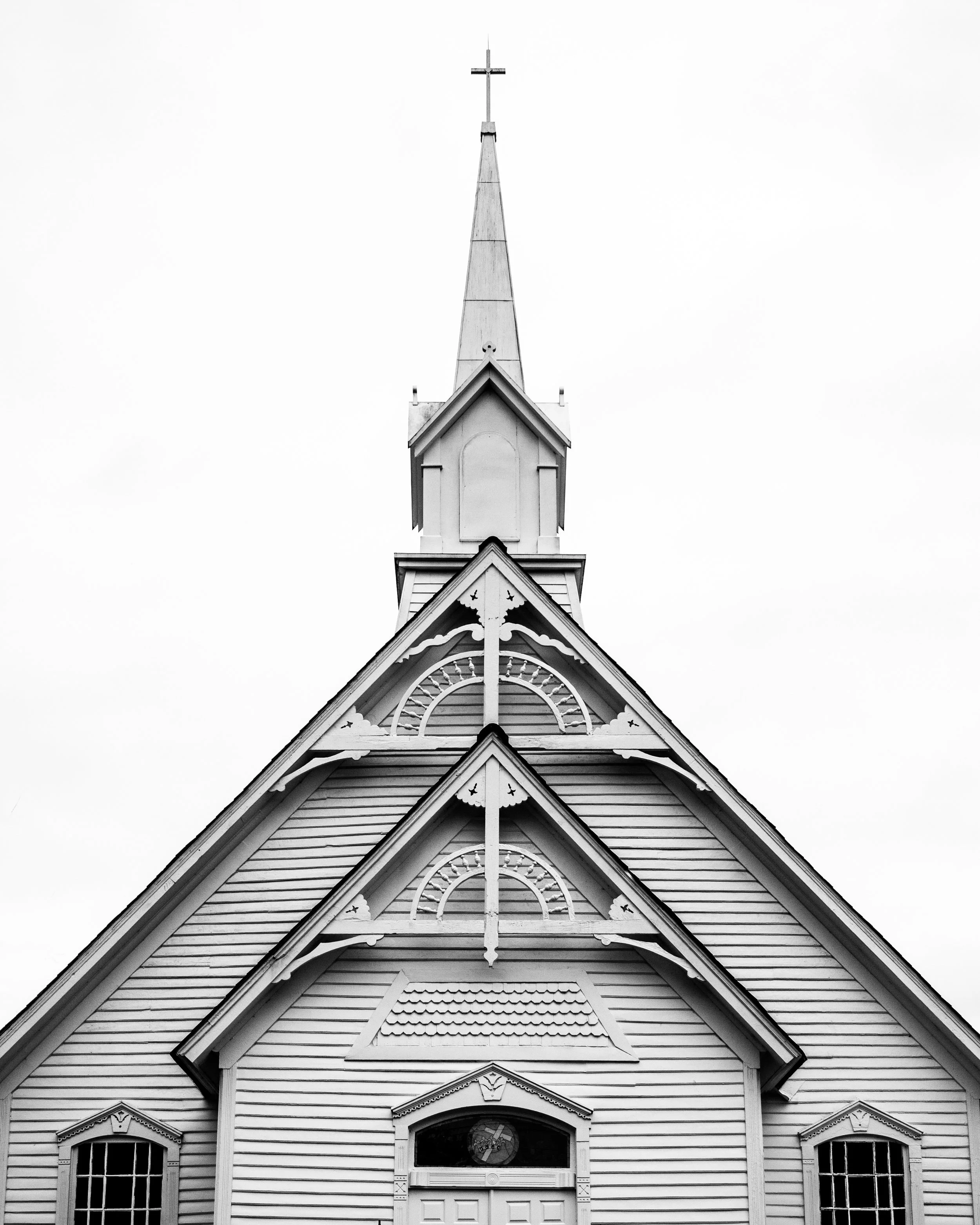 Front view of a white wooden church with a tall steeple and cross at the top, decorative trim, and arched windows.