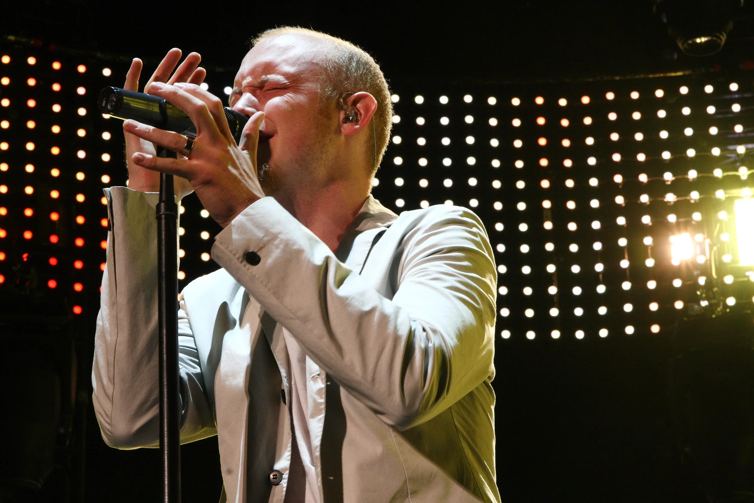 A man singing into a microphone on stage, wearing a white jacket, with bright lights in the background.