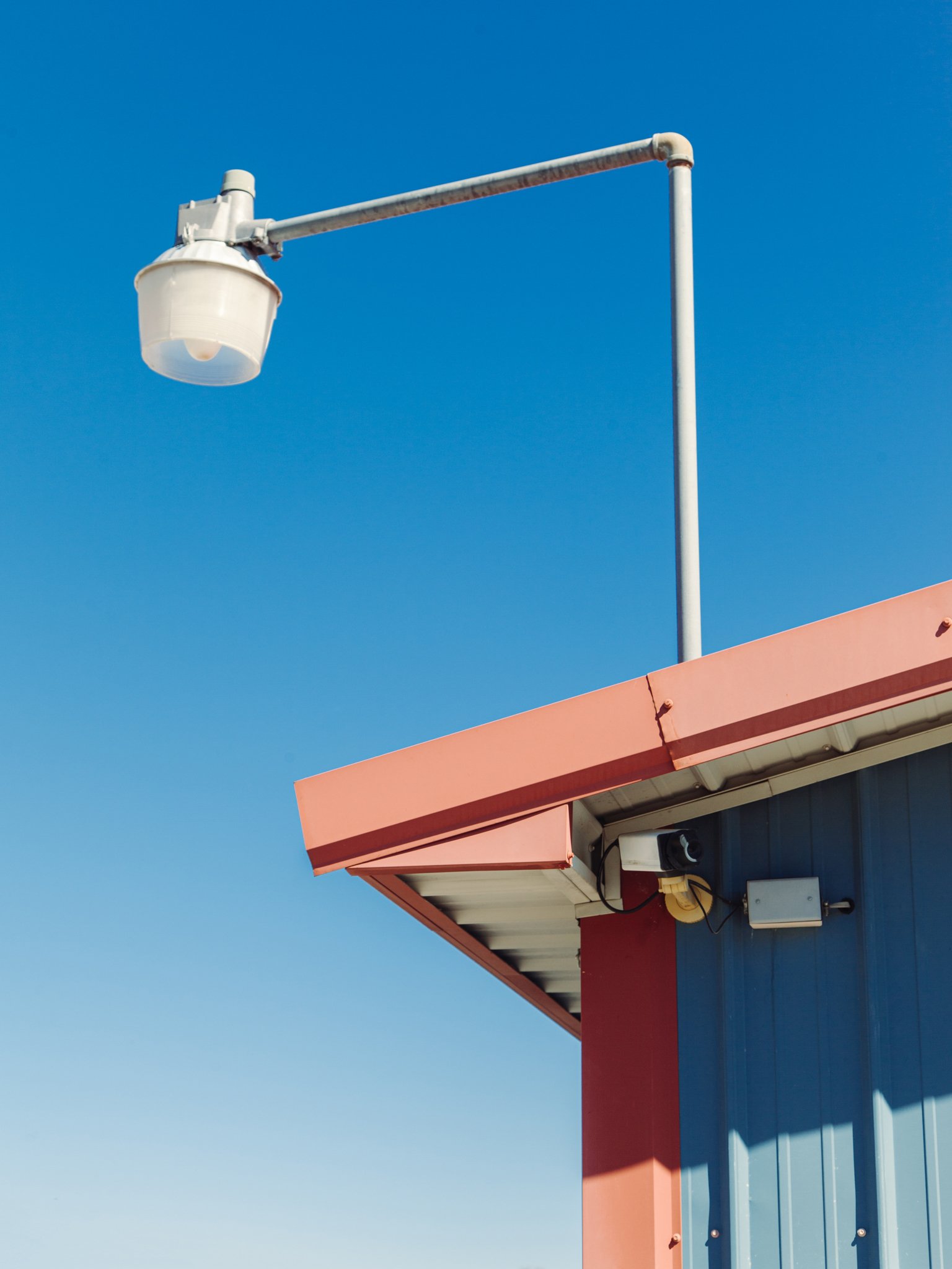 Close-up of a rooftop with an orange trim, blue and gray siding, and a security camera mounted under the eaves, against a clear blue sky.