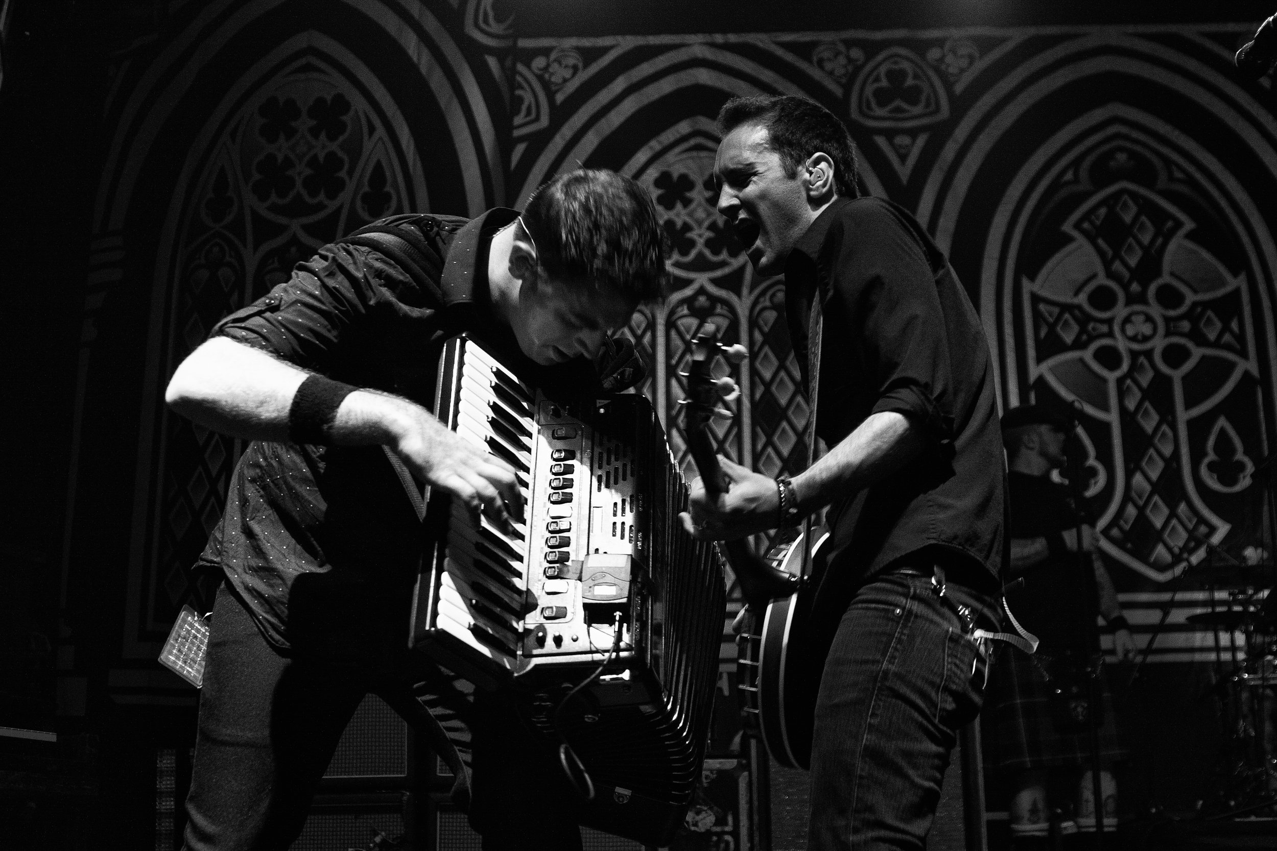 Two musicians perform passionately on stage; one plays an accordion and the other sings or shouts, with a gothic artwork background in black and white.