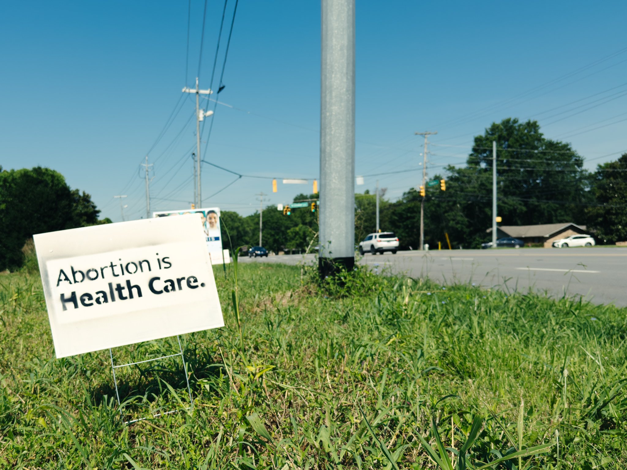 A sign on the grass that reads 'Abortion is Health Care.' Near the sign, there are power lines, trees, and cars on a busy road with traffic lights.