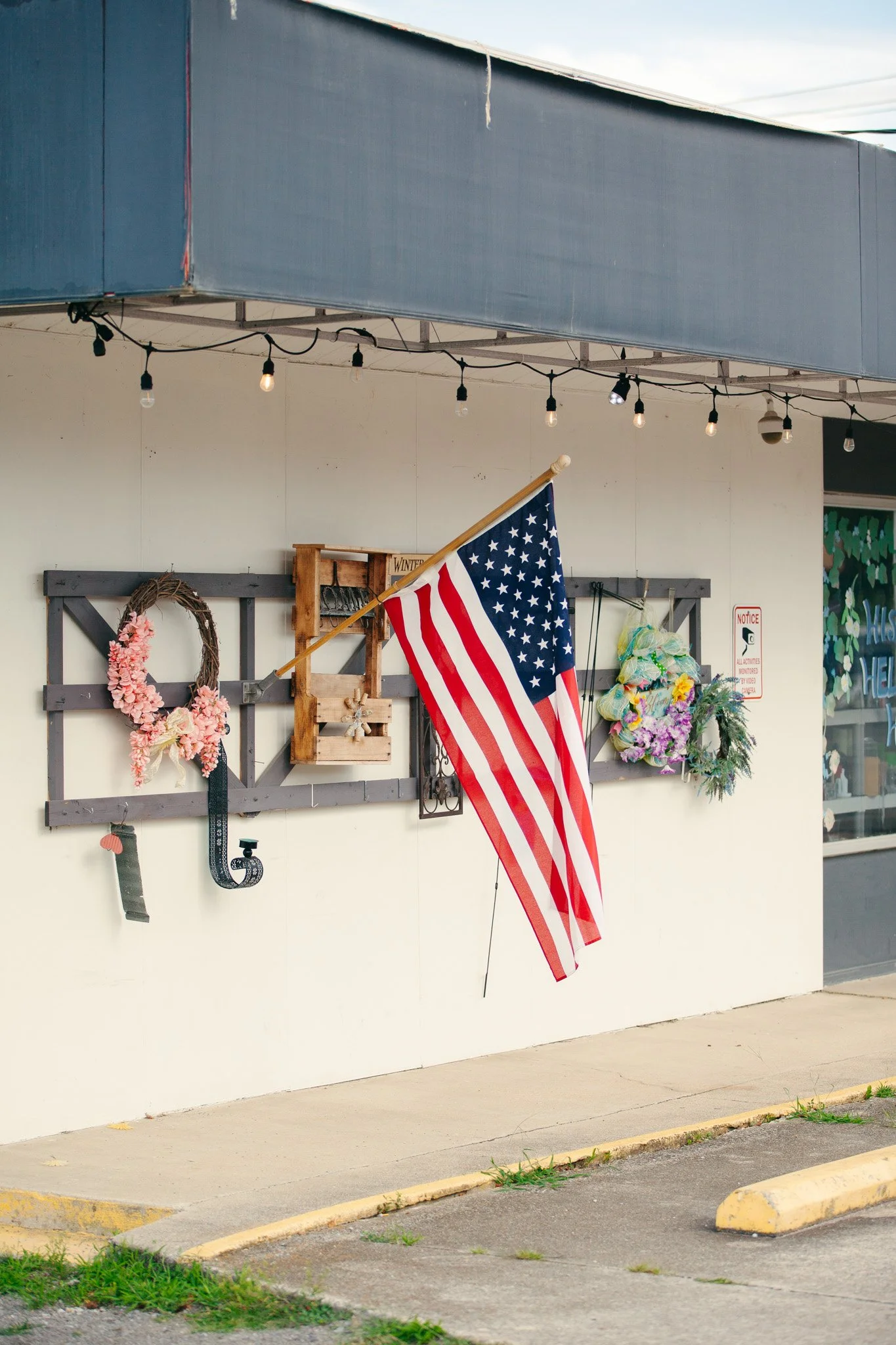 American flag hanging from a wooden post outside a building decorated with floral wreaths and flowers, with string lights above and a sign on the stall.