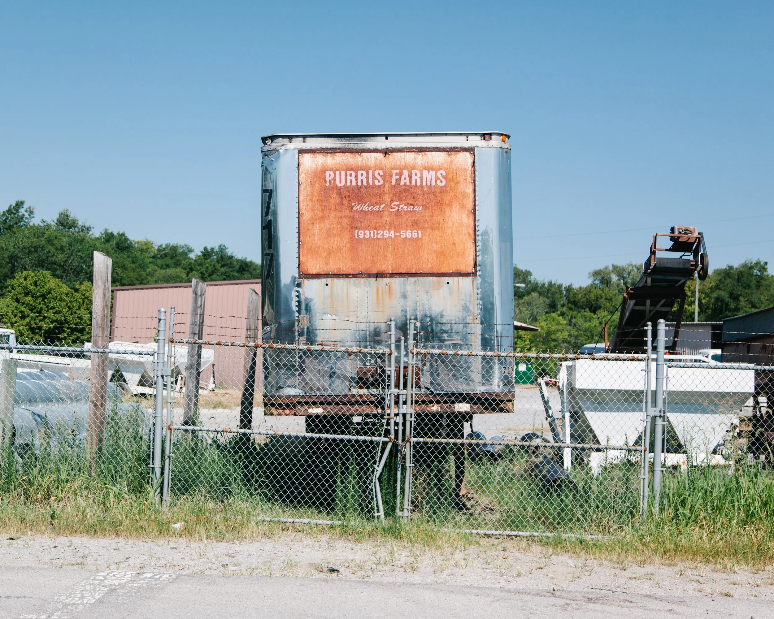A weathered sign on a metal trailer reading 'Purris Farms Wheat Straw' with a phone number, behind a chain-link fence and surrounded by greenery under a clear blue sky.