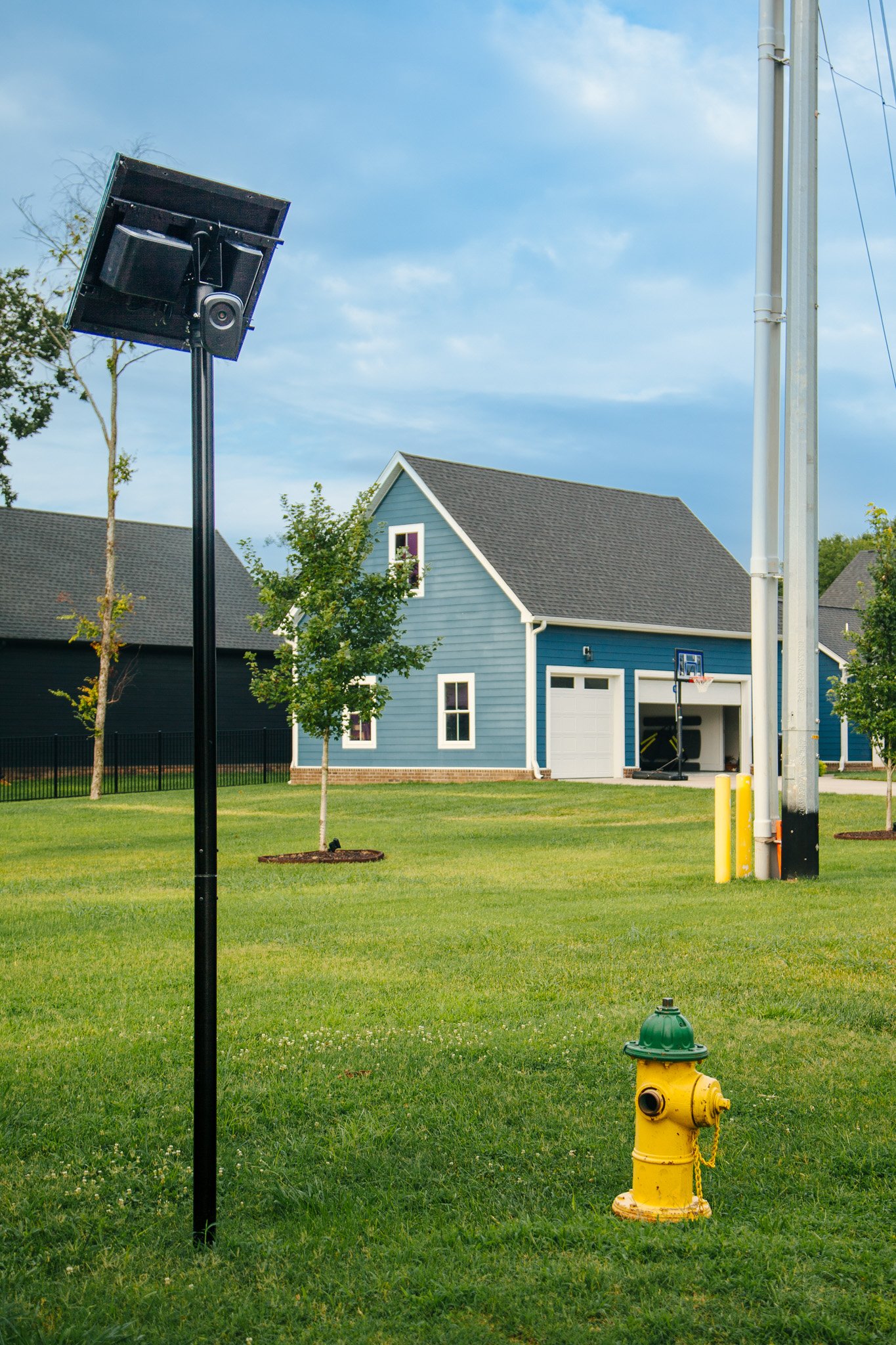 A residential neighborhood with a blue house, a basketball hoop, a fire hydrant, and a flock camera installed on a pole, under a partly cloudy sky.