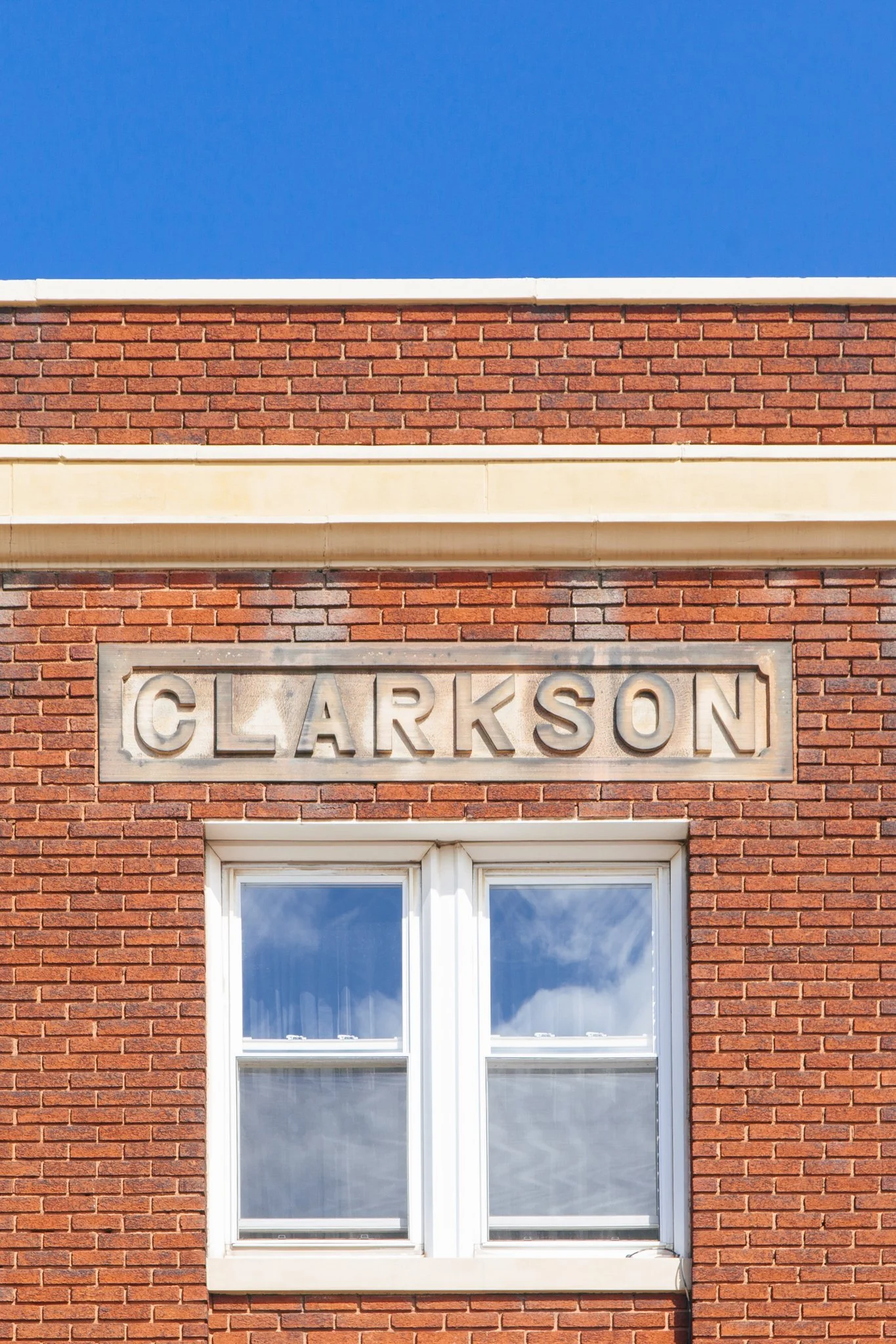 Close-up of a brick building with a white window and a stone sign reading 'Clarkson' above the window. The sky is blue with some clouds reflected in the window.