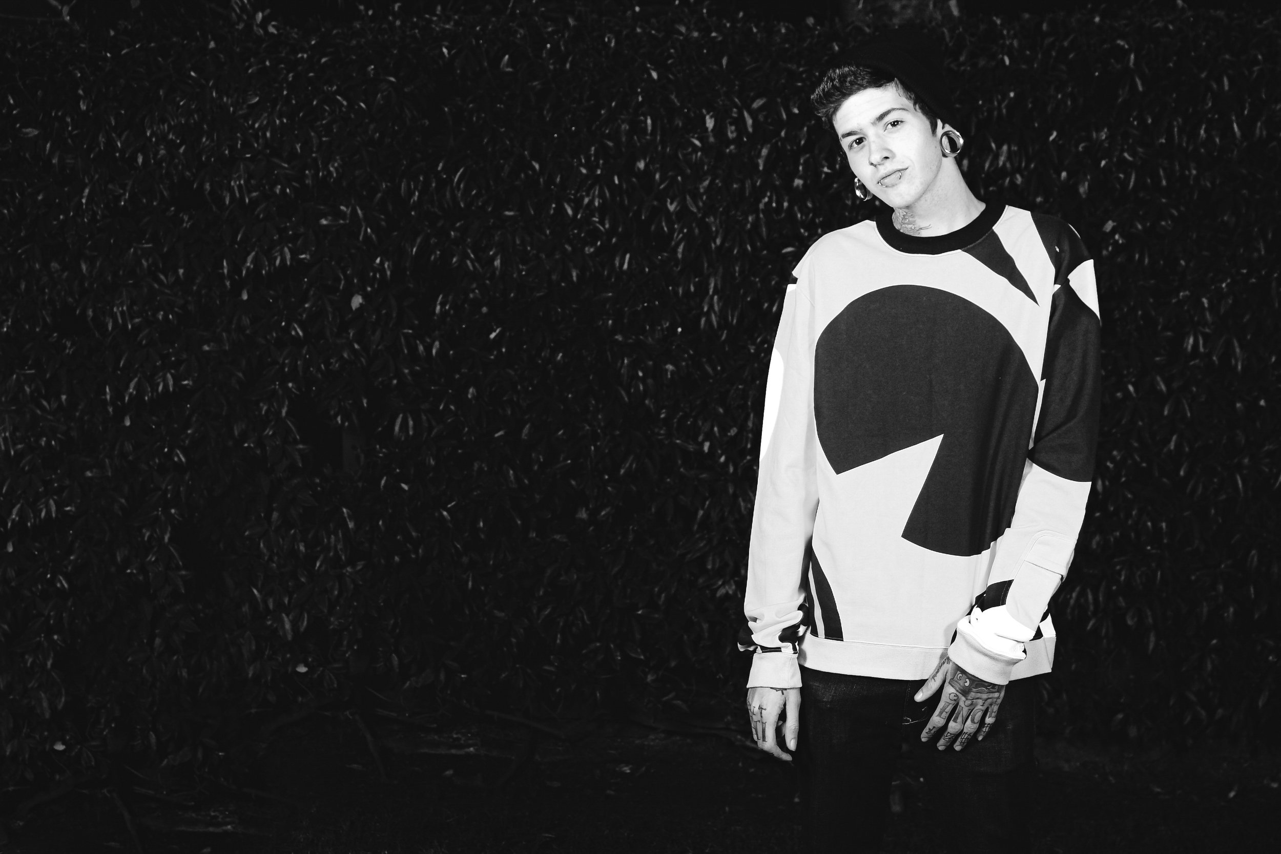 A young man with tattoos, tattoos, large earrings, and ear gauges, standing in front of a dark leafy background, wearing a graphic black and white long-sleeve shirt, with a confident pose.
