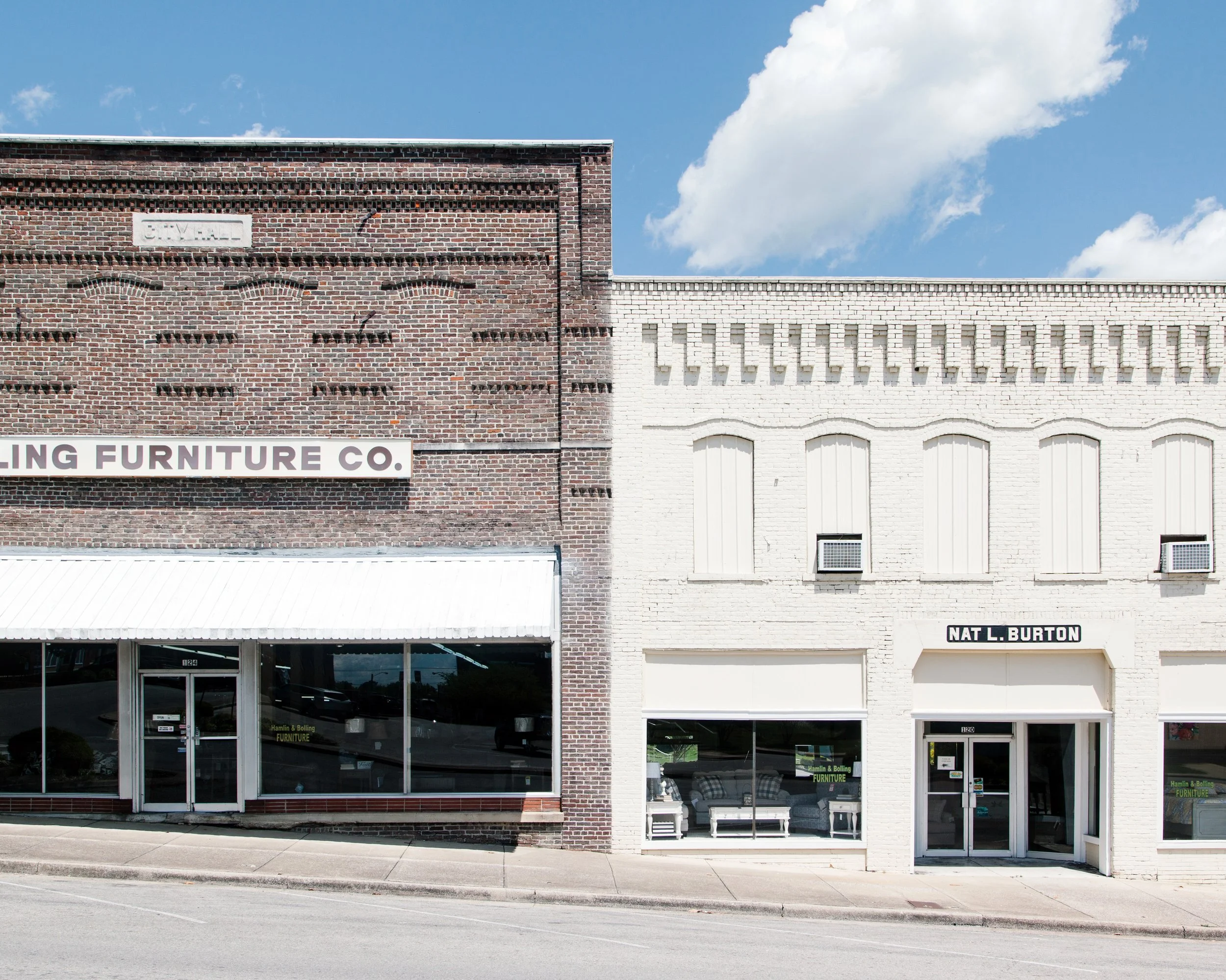 A street view of two adjacent commercial buildings with brick and white facades, displaying large glass windows and doors, with signage indicating furniture stores, under a partly cloudy sky.