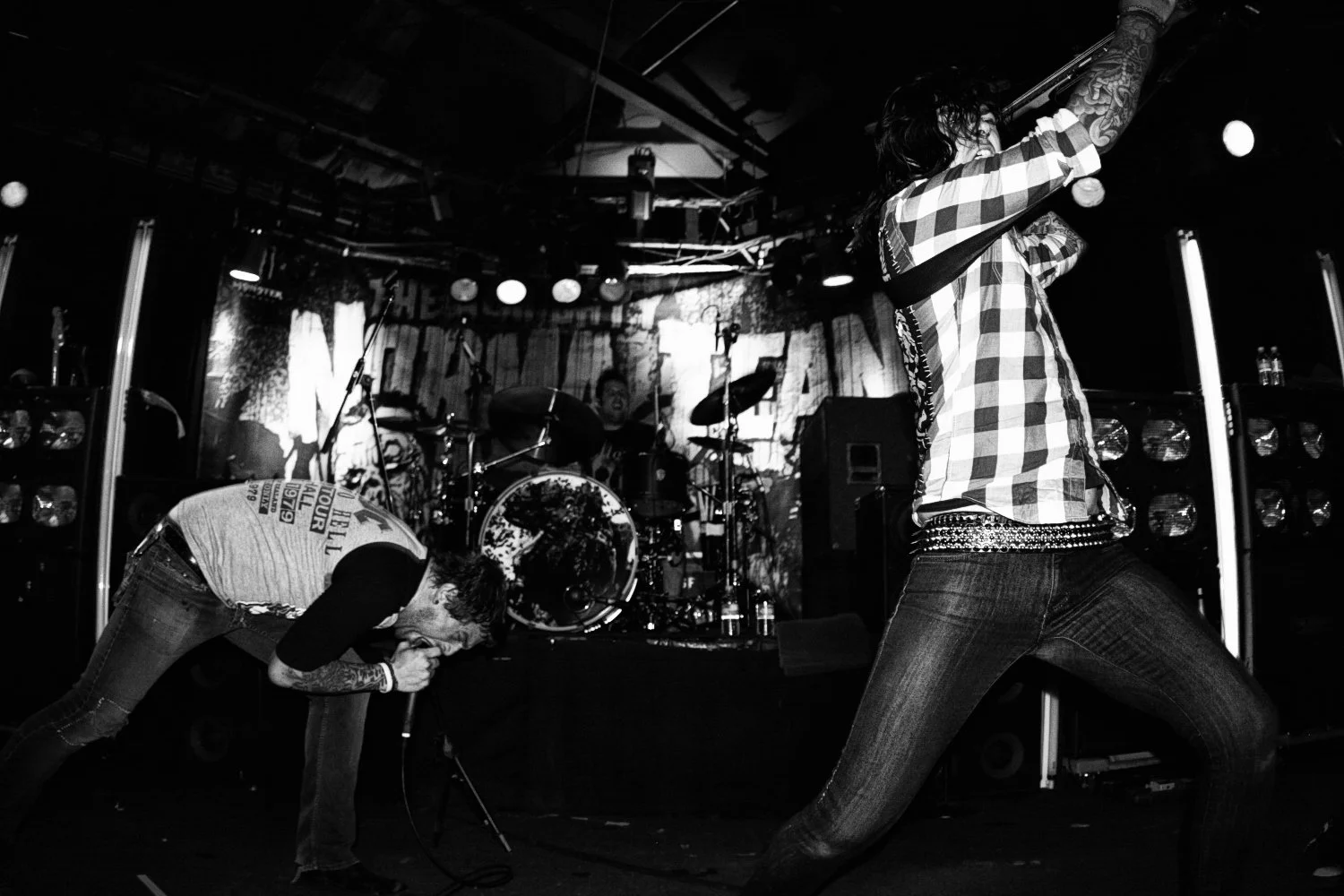 Black and white photo of a rock concert with a singer in a plaid shirt and tattooed arm singing into a microphone, a drummer in the background playing drums, and a second performer bending forward, all on stage with musical equipment and lighting.