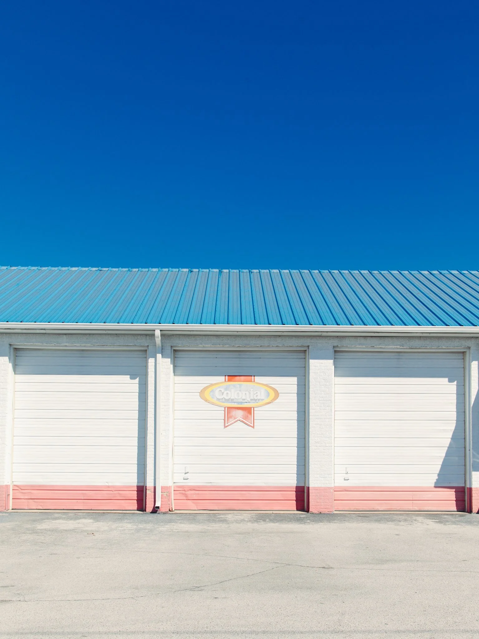 A building with a white brick exterior and a blue metal roof, featuring a sign that reads 'Colonia' and three closed white garage doors beneath the sign. The scene is set against a clear blue sky.