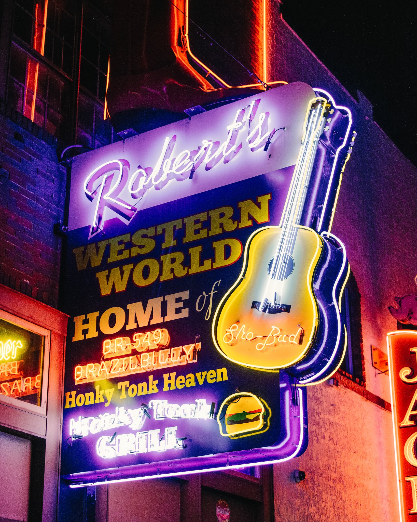 A neon sign for the restaurant 'Robert's Western World' with a brightly lit guitar and a burger graphic, advertising various food items and beverages, brightly colored in purple, yellow, and white neon lights.