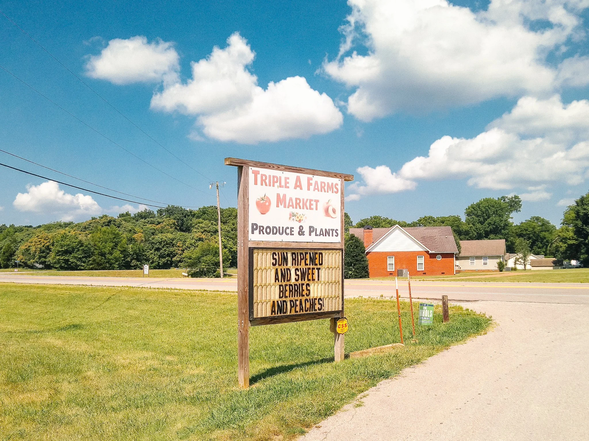 A roadside sign for Triple A Farms Market advertising produce and plants, with a list of sun-ripened, sweet berries and peaches, set against a background of houses, trees, and a blue sky with scattered clouds.
