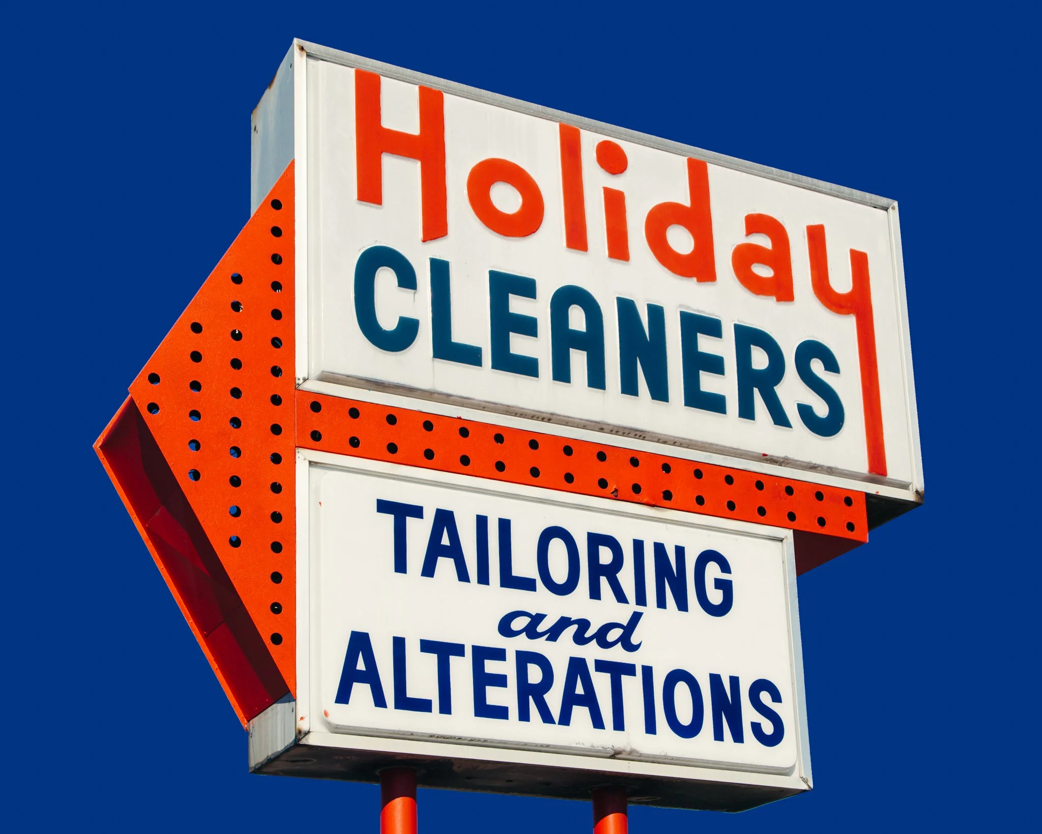 A large illuminated sign with the words 'Holidy Cleaners' in red and blue letters, with a partly visible additional message below, against a clear blue sky.