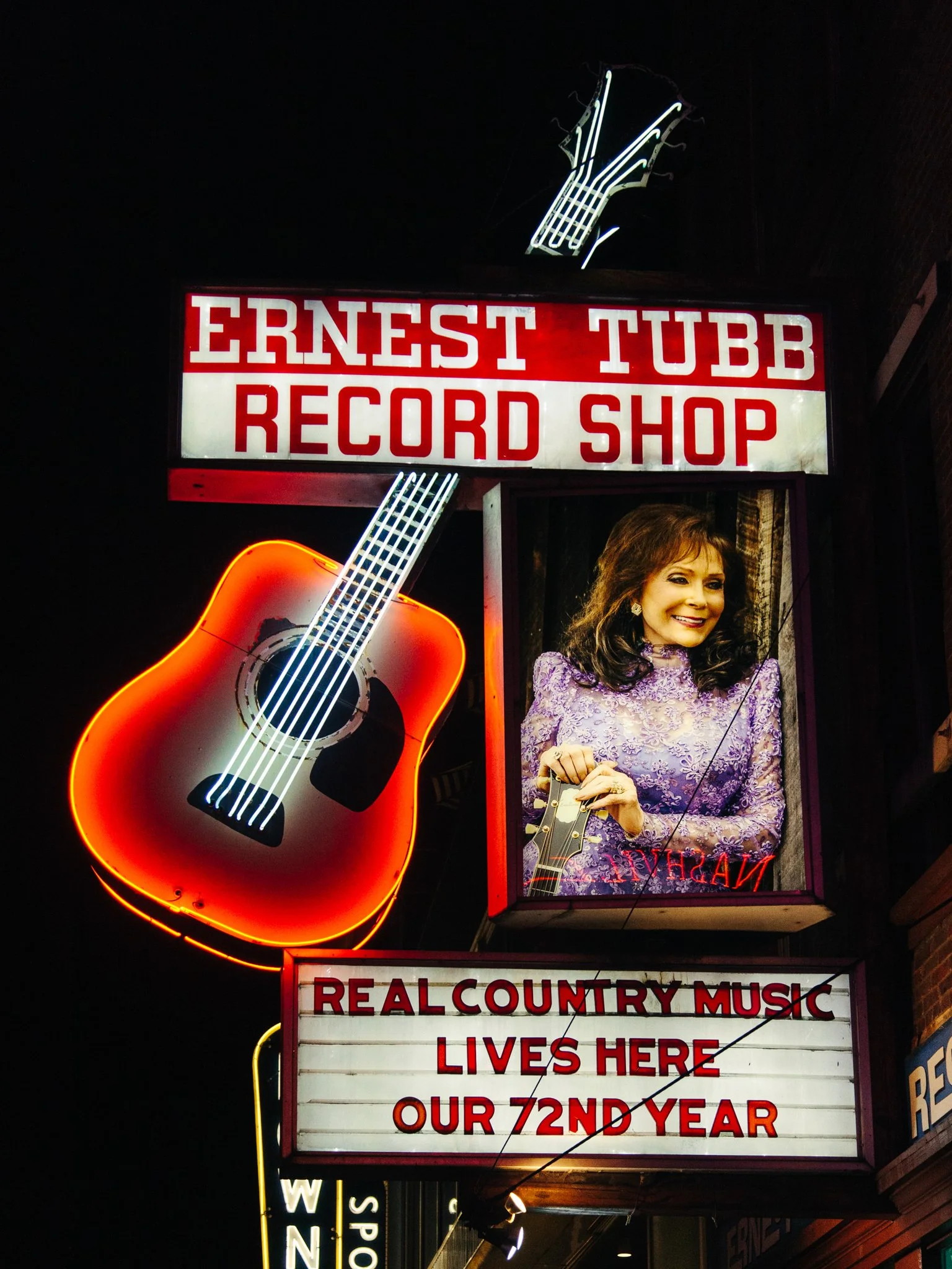 Neon sign and billboard for Ernest Tubb Record Shop, featuring a neon guitar outline and a photo of a woman with dark hair in a purple patterned top, smiling.