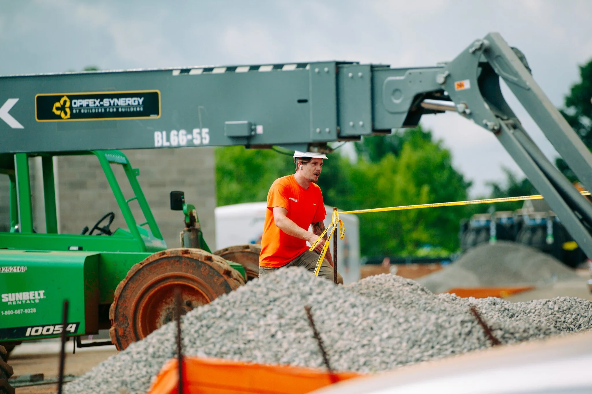 A construction worker in an orange shirt and white cap working on a gravel project at a construction site. There is equipment including a green tractor and a large crane in the background.