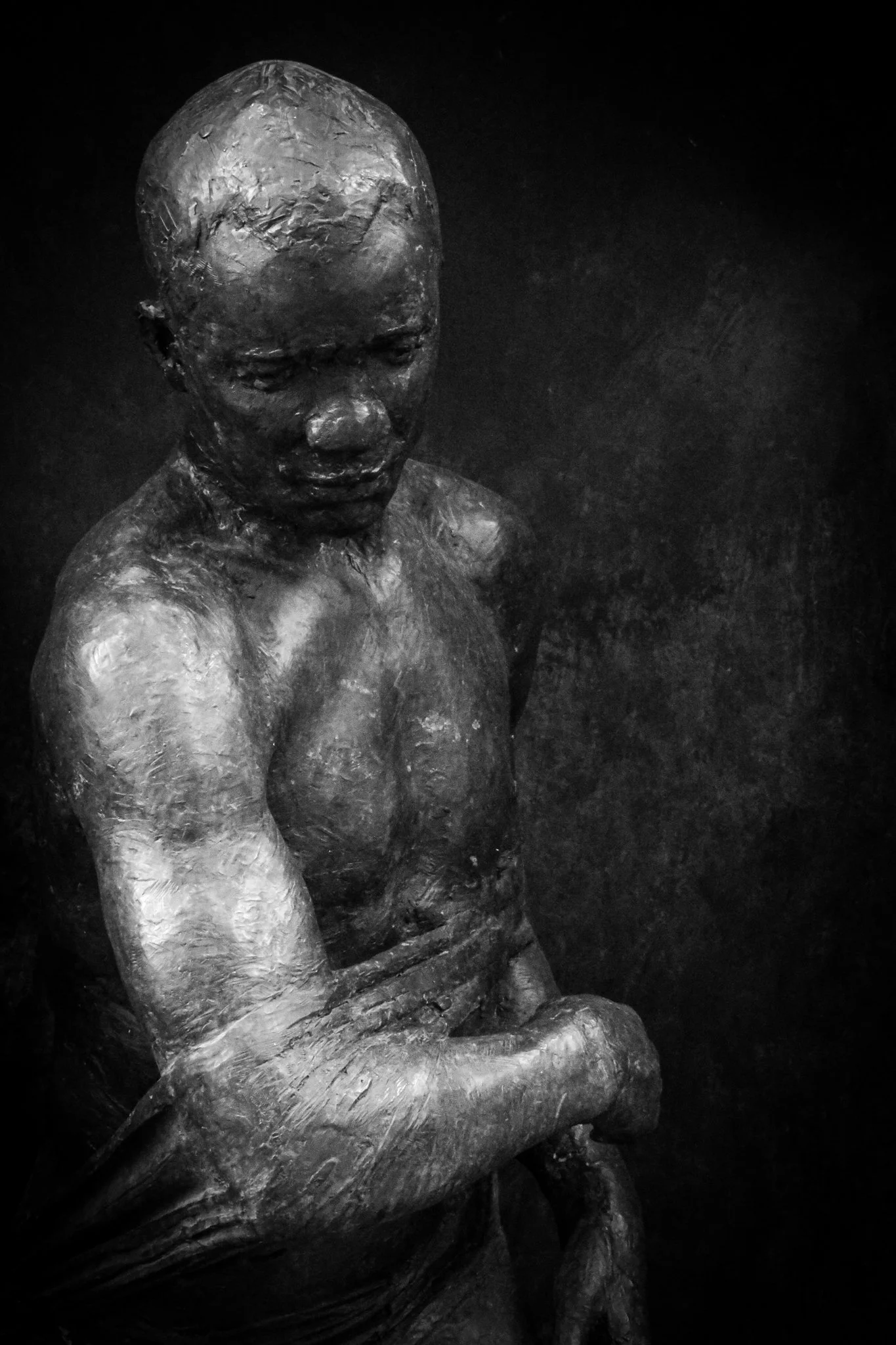 Black and white sculpture of a seated woman with her arms wrapped around her knees, looking downward, against a dark background.